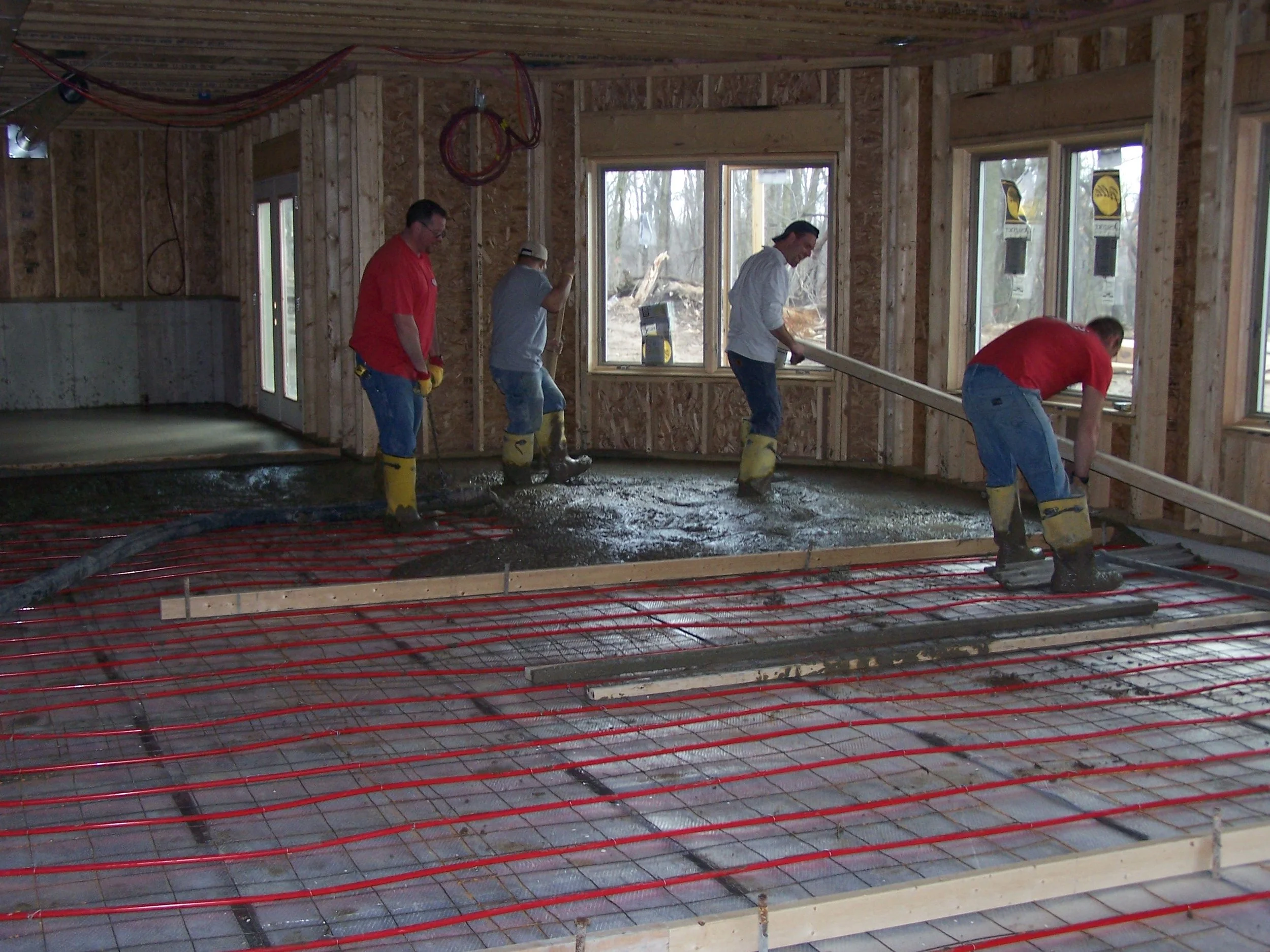 Four workers pouring and spreading concrete inside a house under construction with wooden walls and window frames, with red heating tubing laid on the floor.
