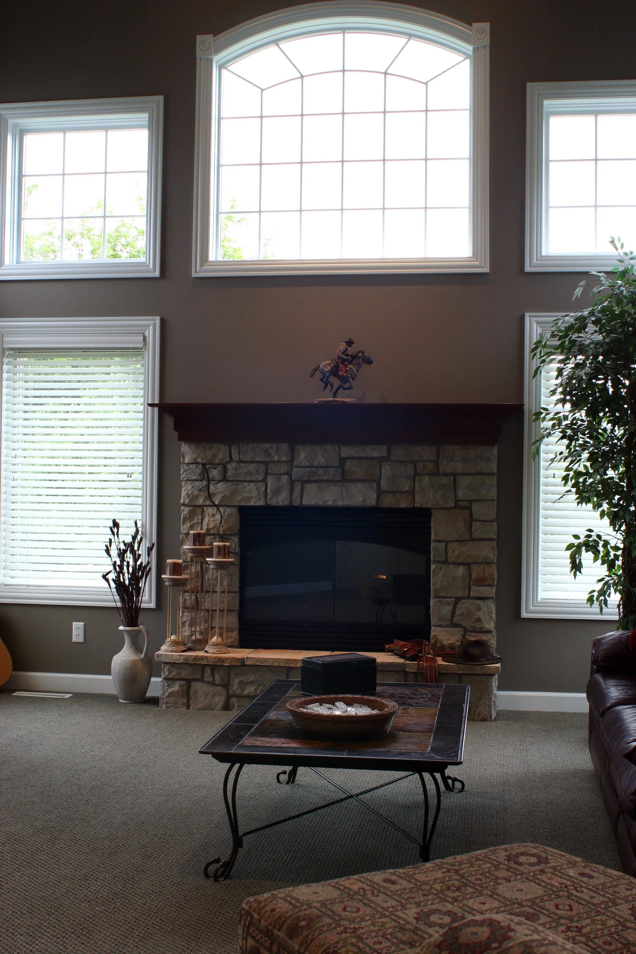 Living room with stone fireplace, large arched and rectangular windows, decorative items on mantle, and a coffee table.