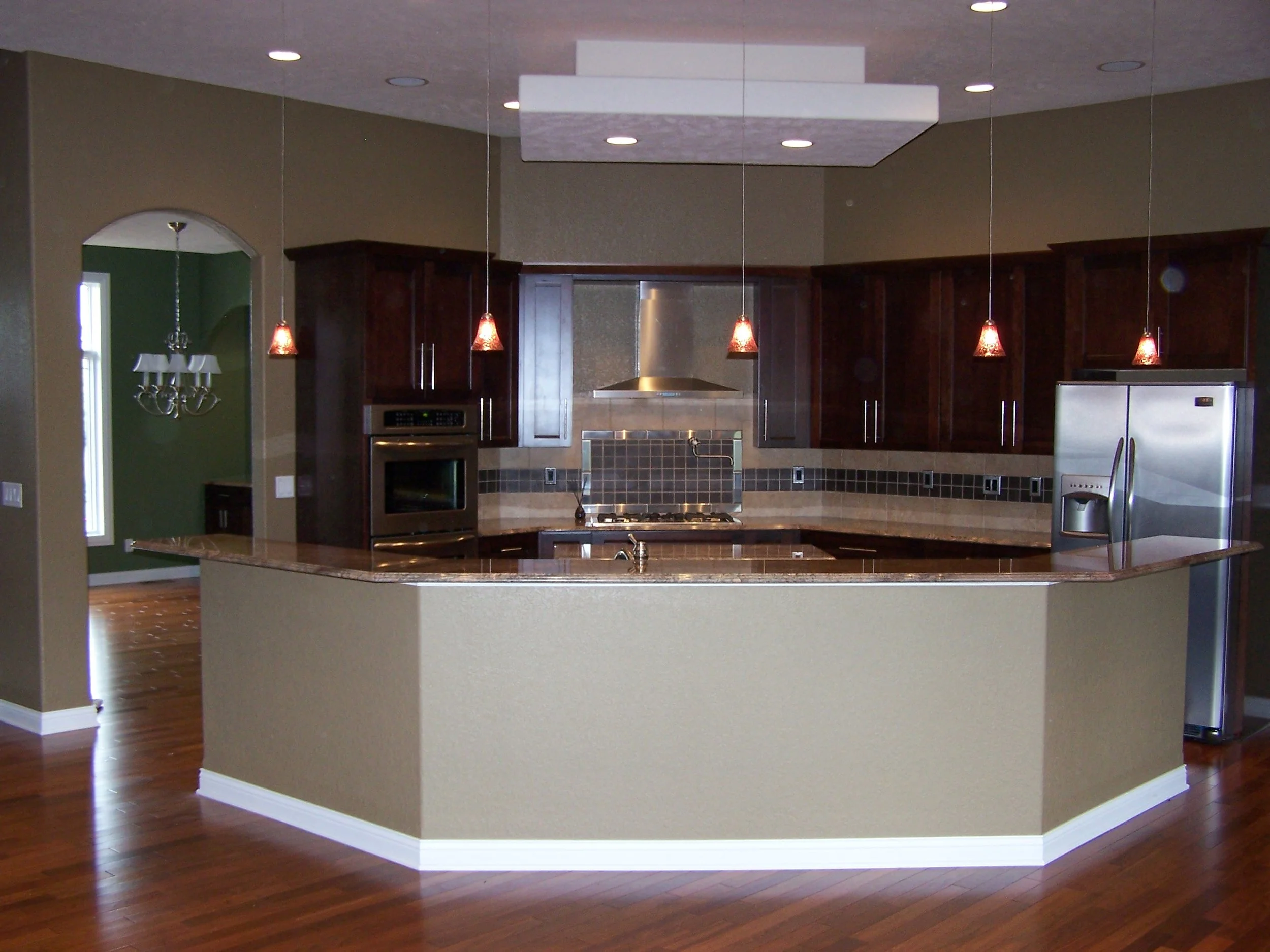 Modern kitchen with dark wood cabinets, stainless steel appliances, and a center island with a brown granite countertop. Pendant lights hang above the island, and a tile backsplash runs along the wall behind the stove.