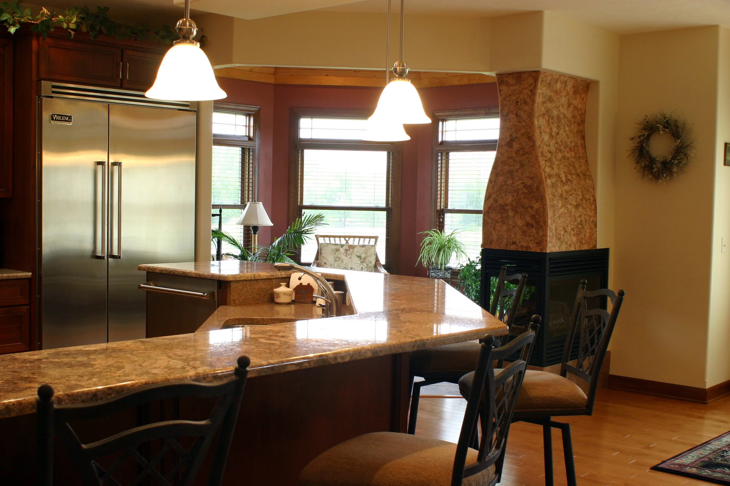 Kitchen with granite countertops, hardwood floors, stainless steel Viking refrigerator, pendant lighting, fireplace, windows with blinds, plants, and chairs at the counter.