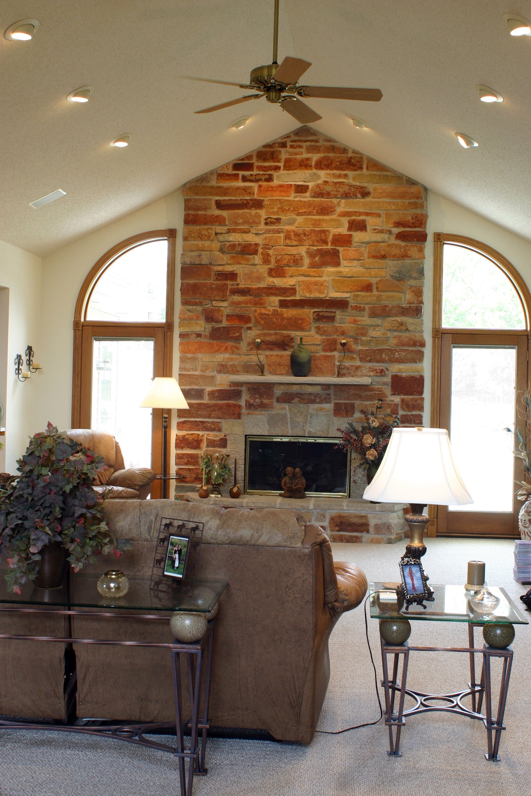 Living room with a large stone fireplace, two arched windows, beige carpet, and furniture including a leather couch and glass tables with lamps and framed photos.