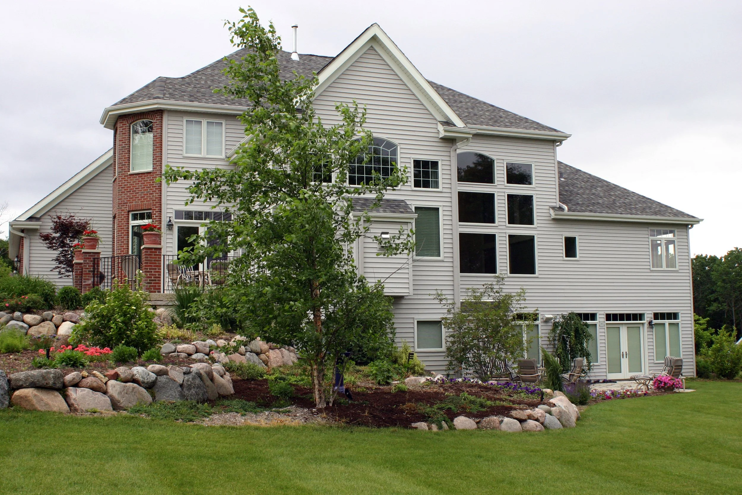 Large multi-story house with a landscaped yard and a small patio in the front. The house has beige siding with white trim, and a brick chimney. There are several windows, including a bay window and a glass door leading outside, with chairs and potted