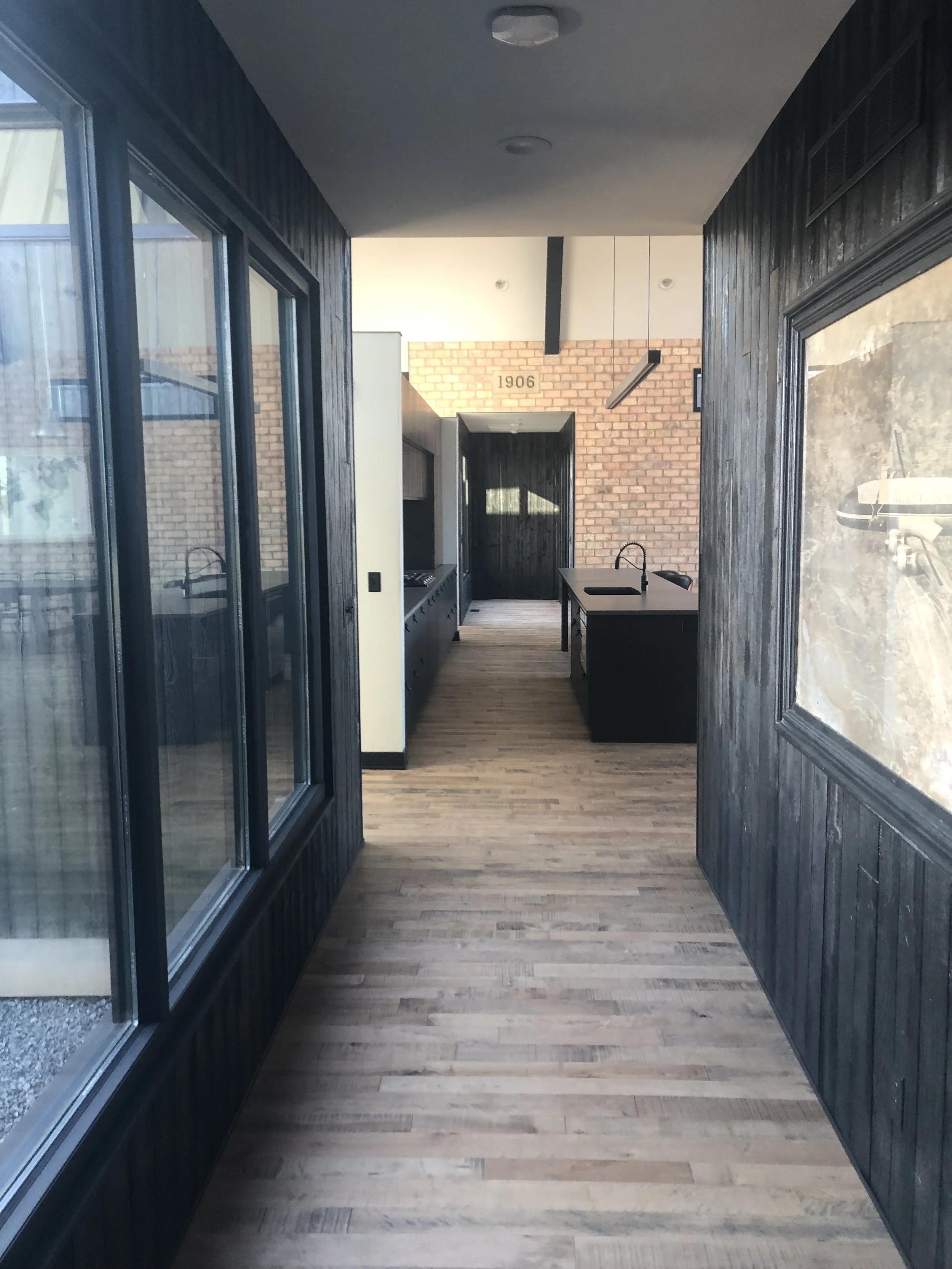 View of a modern kitchen through a hallway with wooden floor and black wooden wall paneling, featuring a brick wall and kitchen island with double sink in the background.