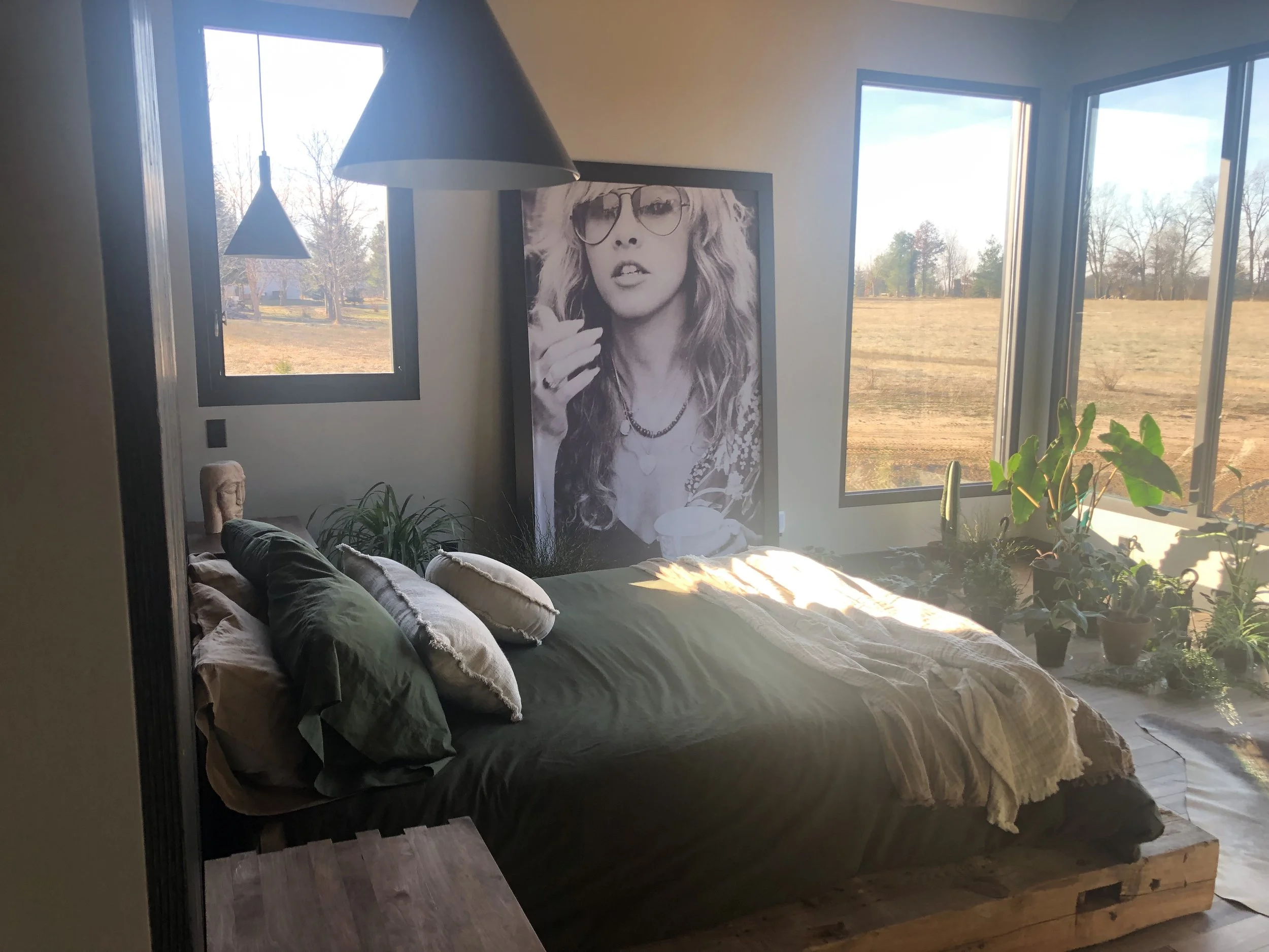 A bedroom with large windows showing a field and trees outside, a bed made with pillows and a blanket, a large black and white portrait of a woman with glasses resting against the wall, and various potted plants near the window.