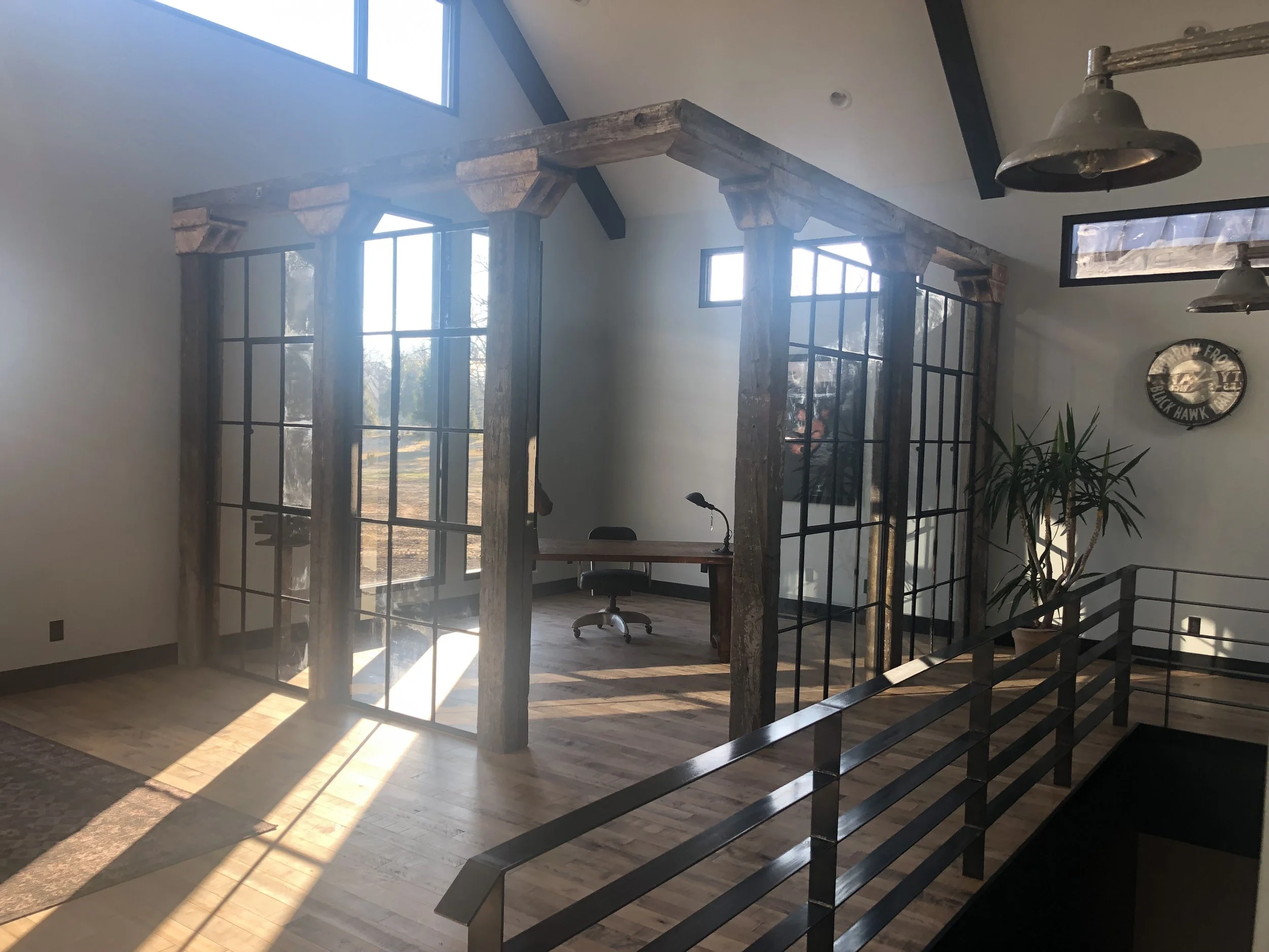 An office space with a rustic wood and black metal enclosed conference area, a desk with a chair inside, large windows, a potted plant, and industrial-style hanging lights.