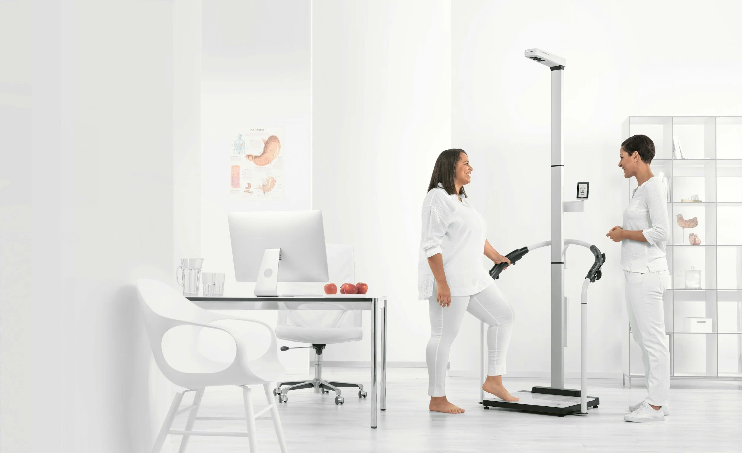 Two women in a white medical or scientific setting, one is standing on a scale with her foot raised, smiling at the other woman, who is holding a device and documenting her weight and health data.