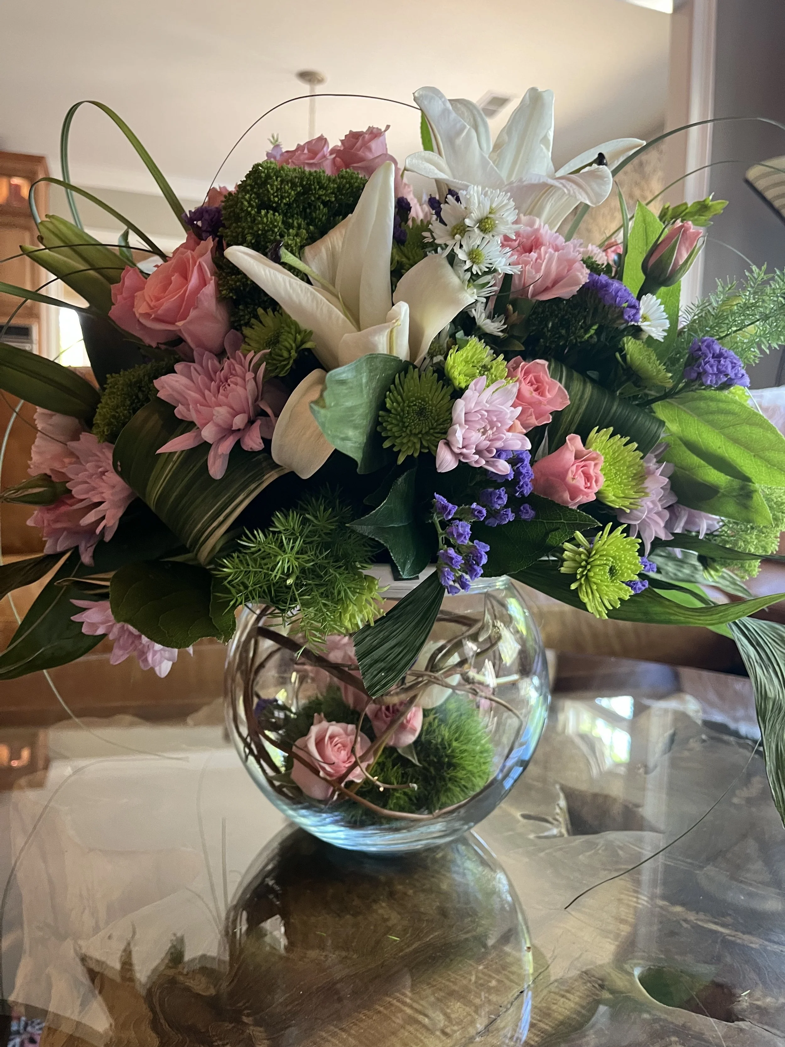 A bouquet of mixed flowers, including white lilies, pink roses, purple statice, green chrysanthemums, and white daisies, arranged in a clear glass vase on a wooden table.