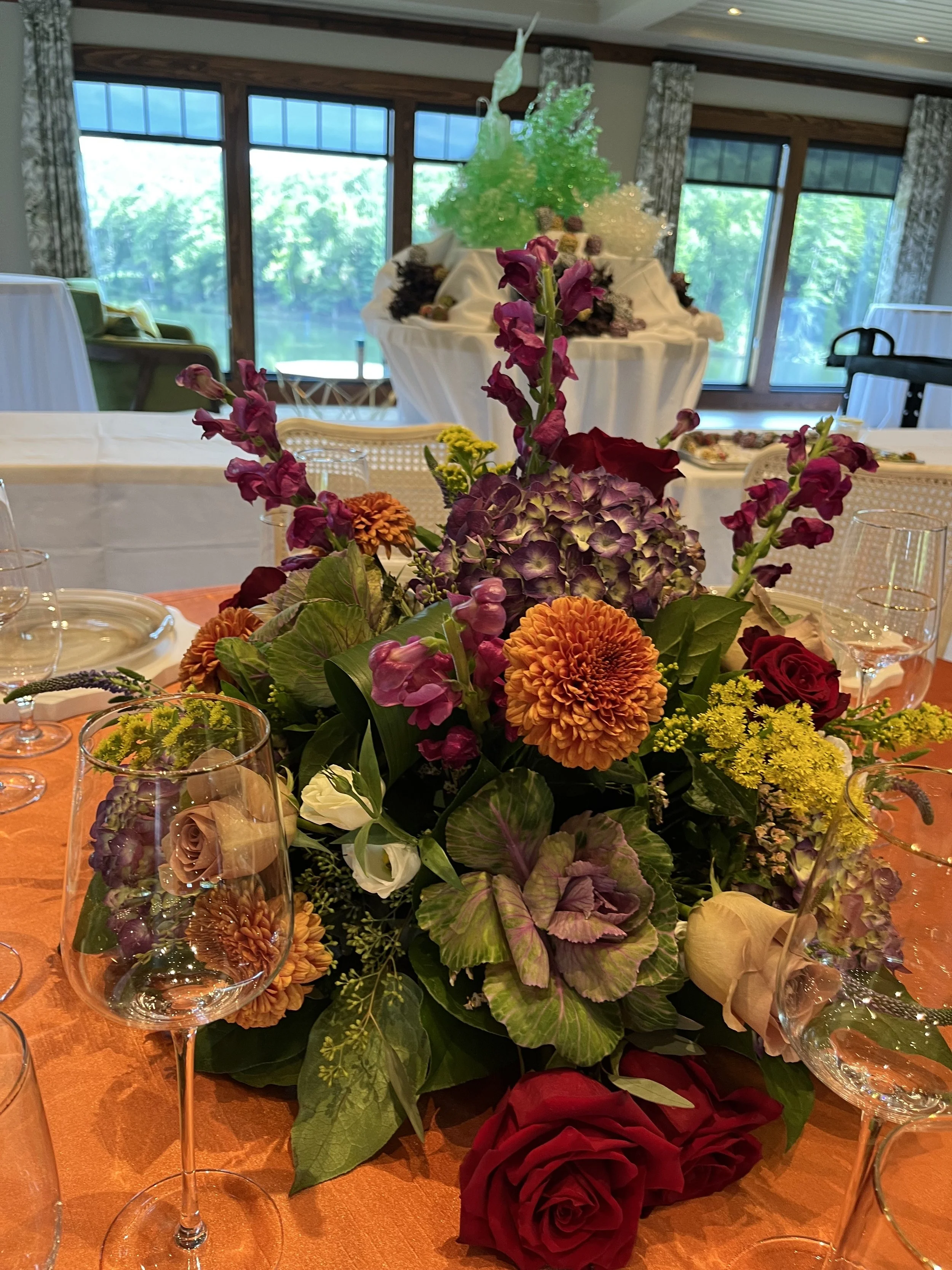 A table centerpiece with a colorful arrangement of flowers, including roses, hydrangeas, and other blooms, is set on an orange tablecloth with empty wine glasses around it. In the background, there are large windows revealing a green outdoor scene.