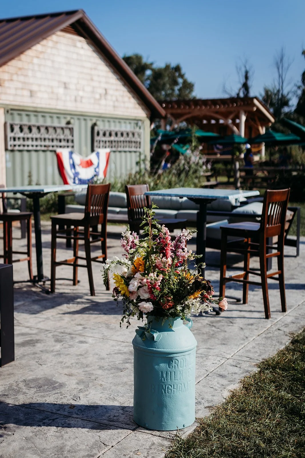 A bouquet of colorful flowers in a light blue milk can with the words 'GROVE MILK BENGHAM' on it, set on an outdoor patio with wooden chairs, tables, and a barn with patriotic bunting in the background.