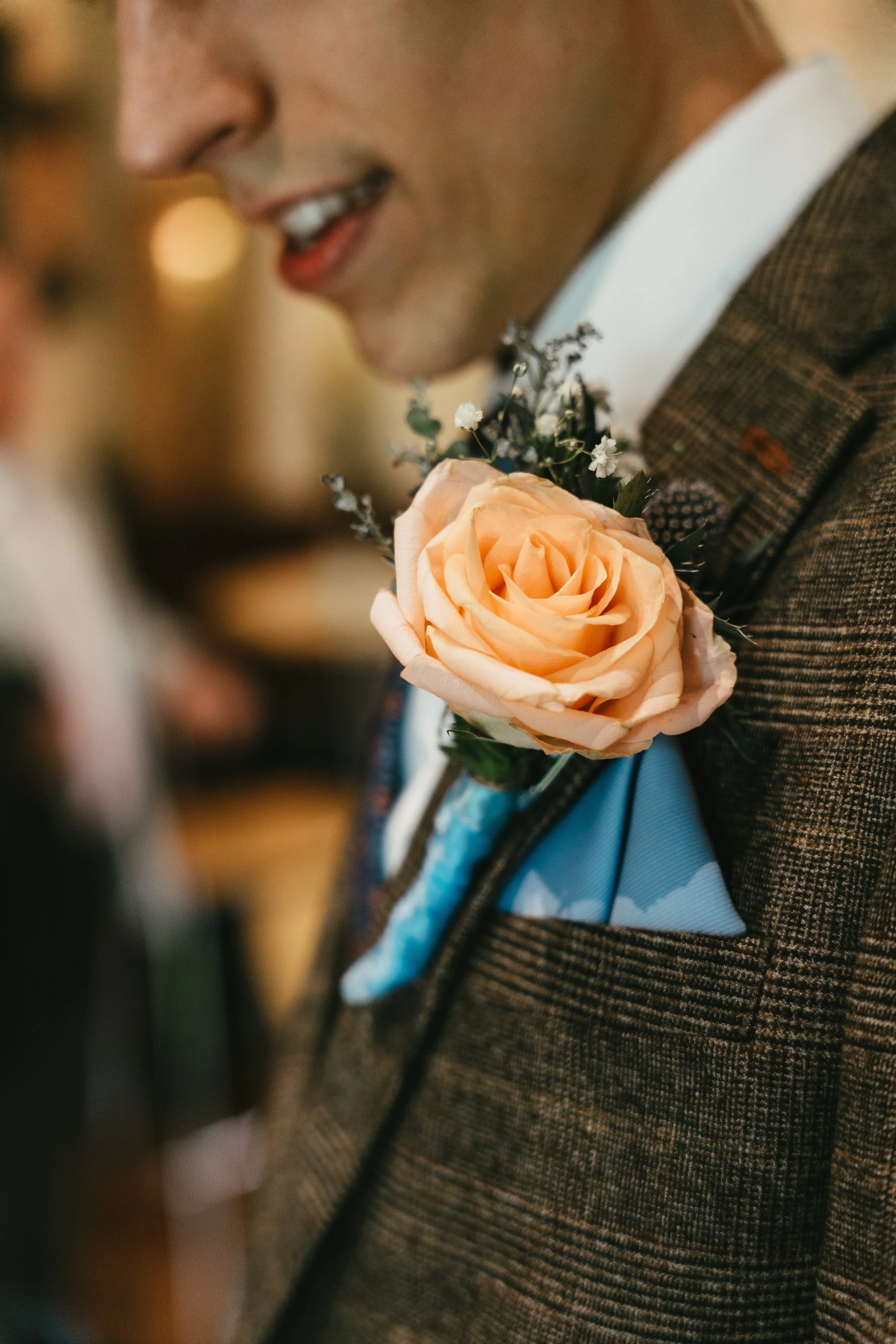 Close-up of a man in a brown checked suit with a peach-colored rose boutonniere on his lapel and a light blue pocket square.