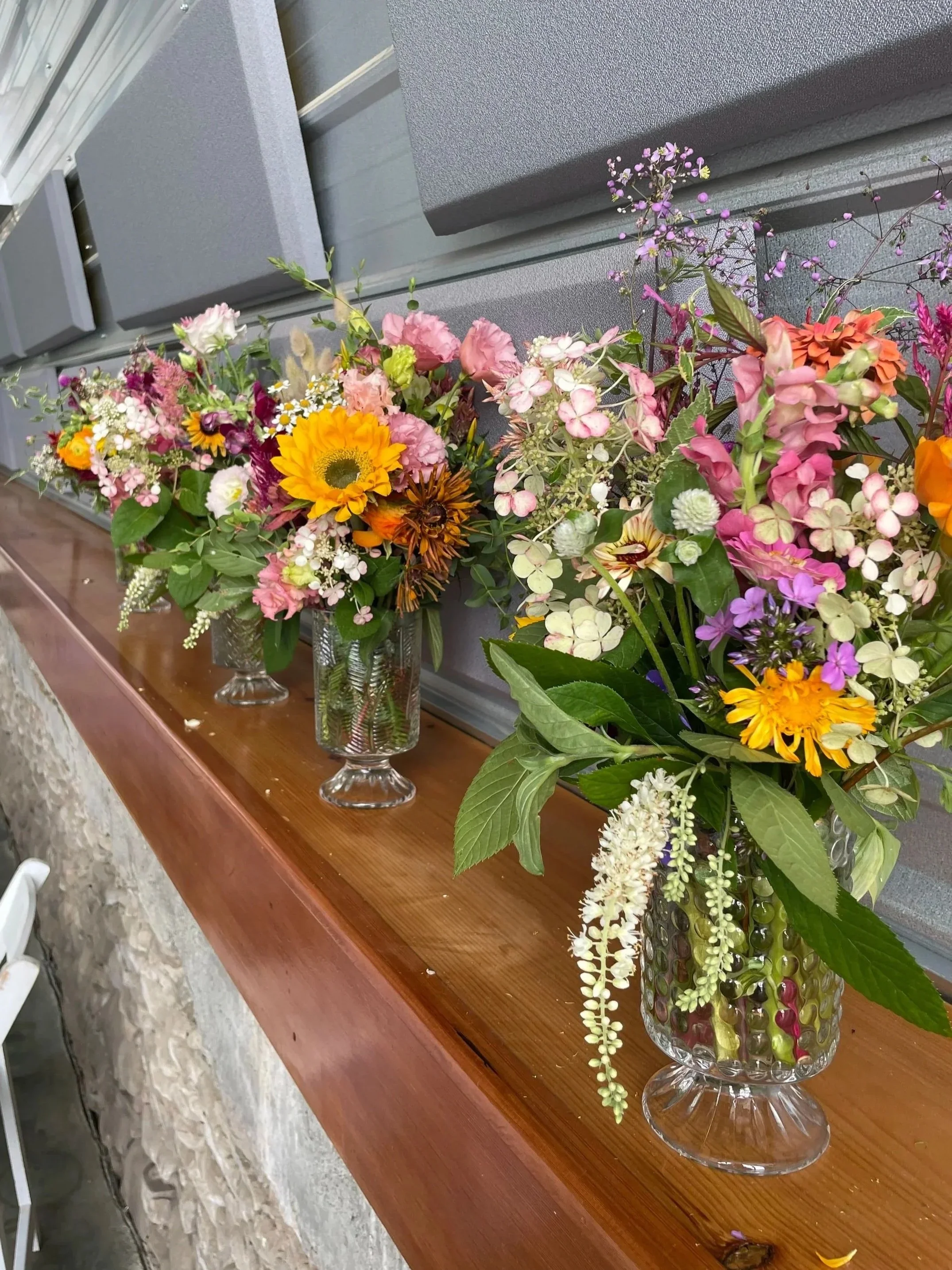 Three glass vases filled with colorful flower arrangements on a wooden ledge, against a gray wall.