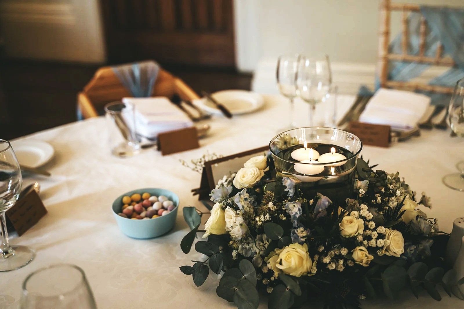 A dining table decorated with a floral centerpiece of white roses, baby's breath, and greenery, with floating candles in a glass bowl on top, surrounded by wine glasses, napkins, and small name cards, likely set for a celebration or event.