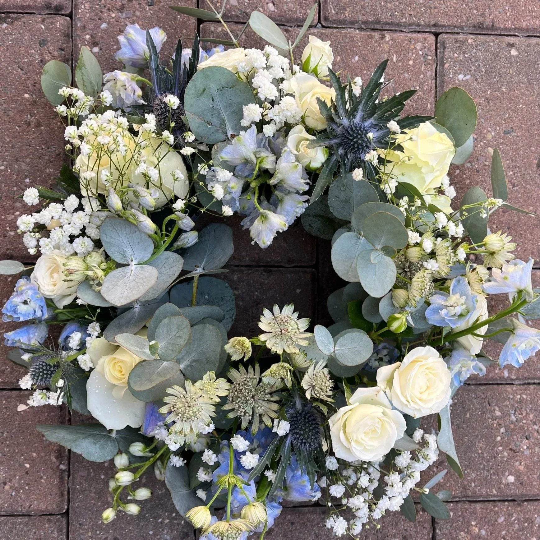 A floral wreath with white roses, blue flowers, eucalyptus leaves, and small white blossoms, laid on brick pavement.
