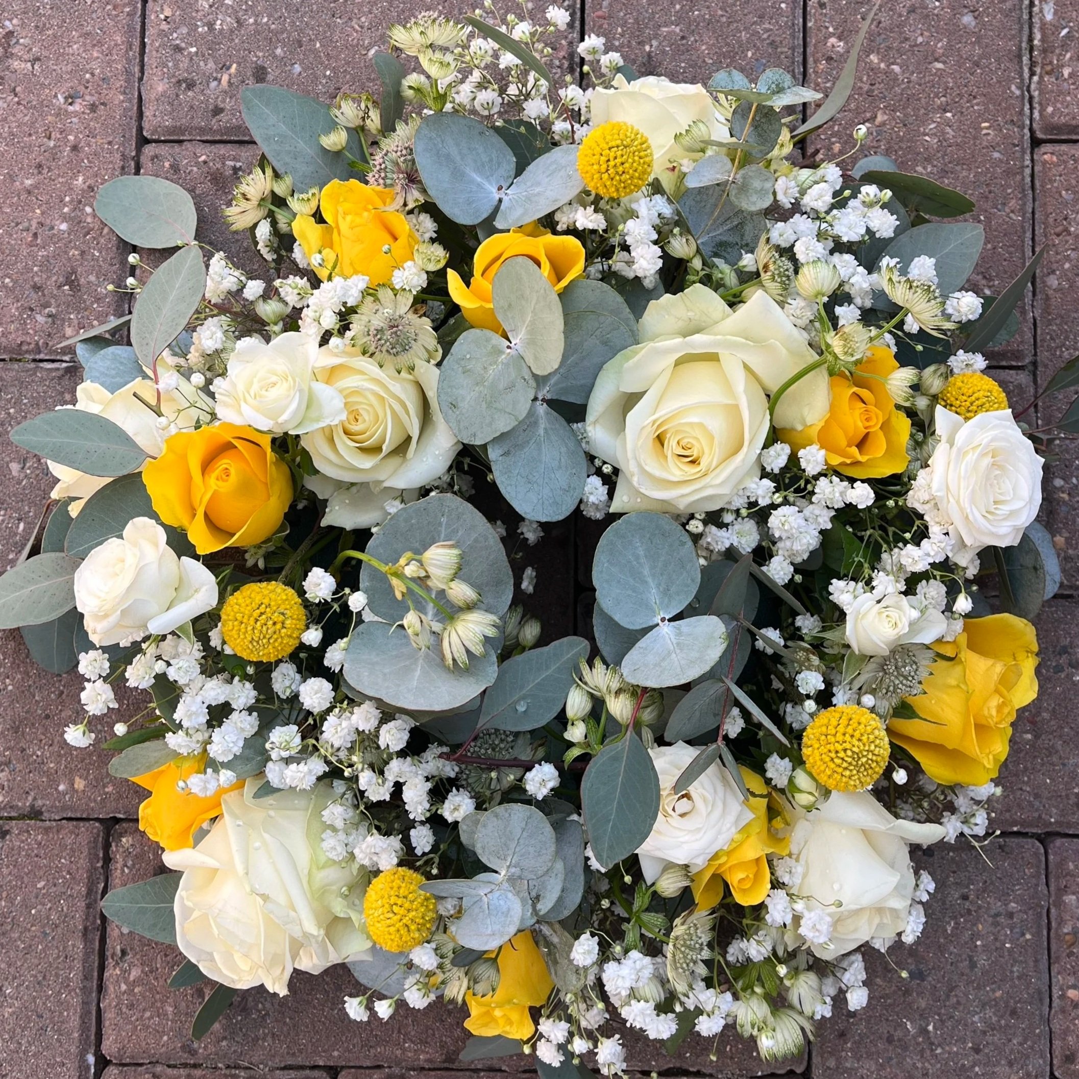 A floral arrangement with white and yellow roses, small white baby's breath flowers, green eucalyptus leaves, and yellow spherical flowers, placed on a brick surface.