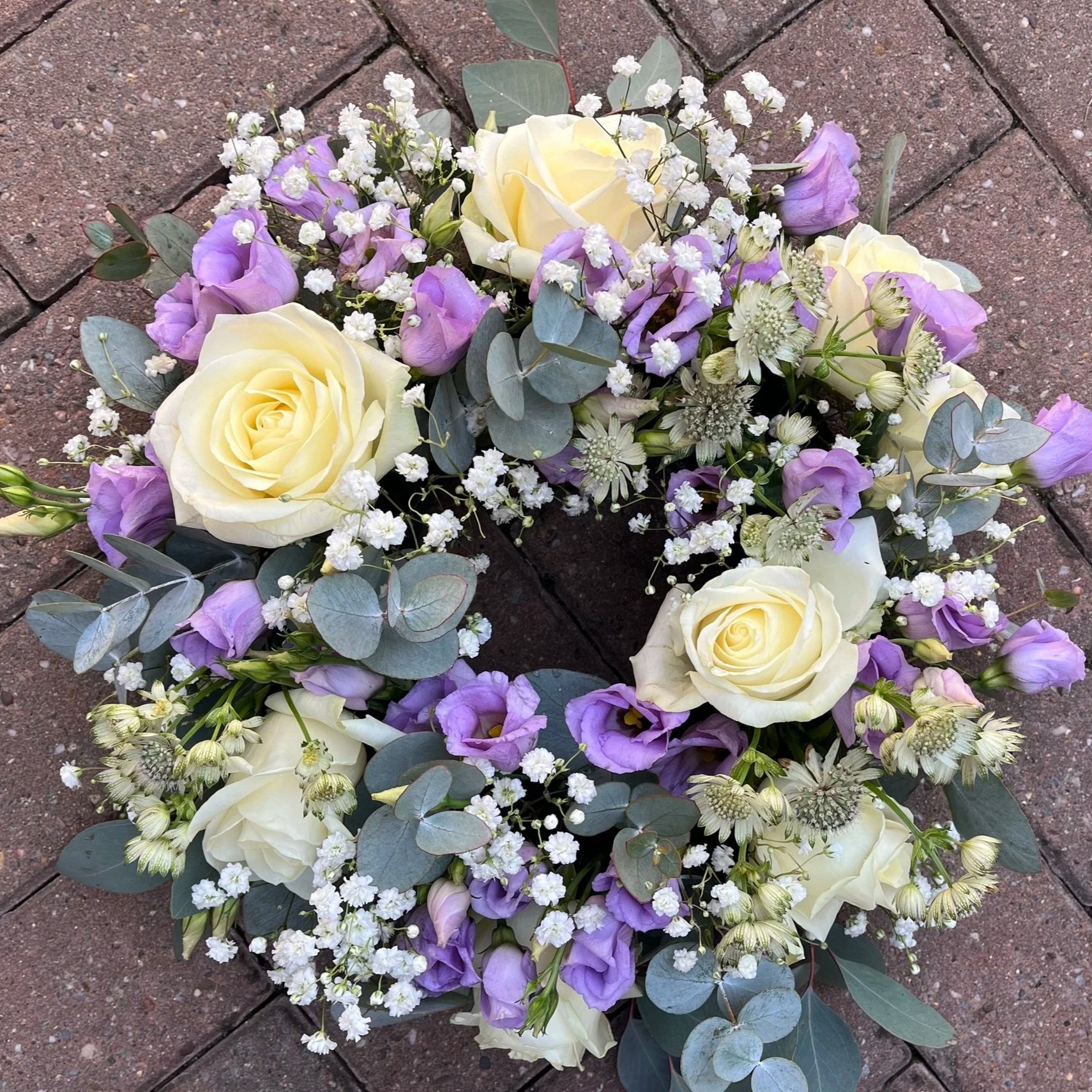 A floral arrangement with white roses, purple lisianthus, baby's breath, and eucalyptus leaves on a brick surface.