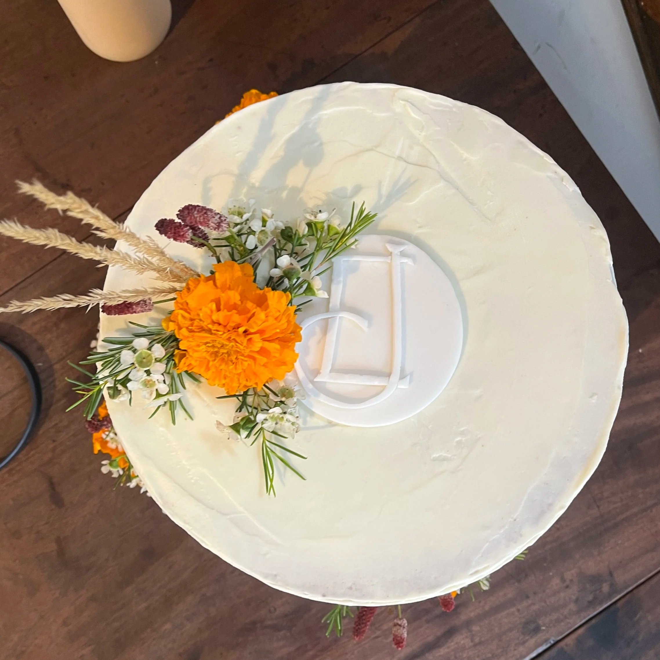 Top-down view of a white frosted cake decorated with orange, white, and purple flowers and greenery, with a monogrammed 'D' in the center.
