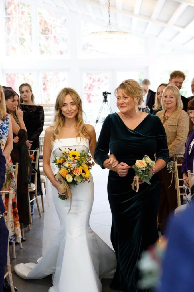 A bride in a white wedding gown holding a bouquet of sunflowers and roses walks down the aisle, accompanied by a woman in a black dress holding a smaller bouquet, during a wedding ceremony with guests in the background.