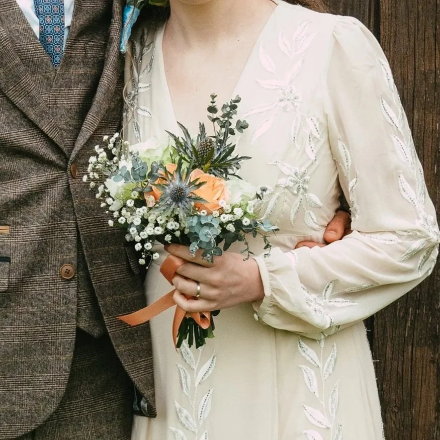 A woman in a white embroidered dress holding a bouquet of flowers, standing next to a man in a brown checkered suit with a blue patterned tie. The woman is wearing a ring on her left hand.