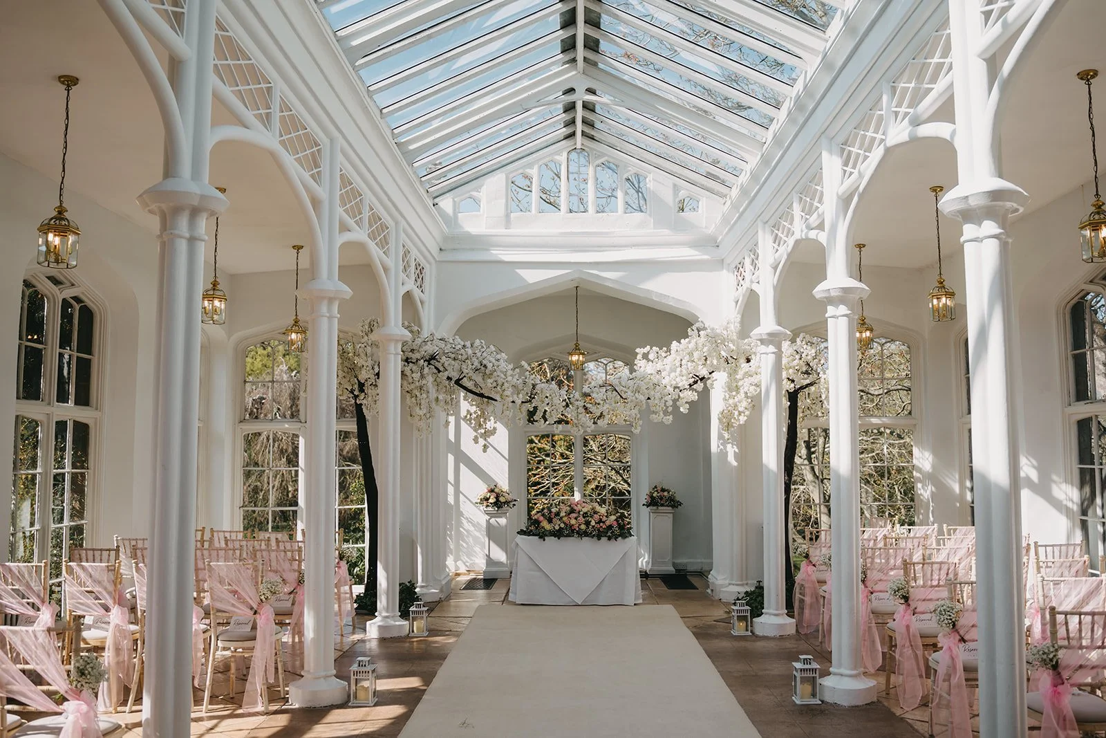 A bright, white enclosed outdoor wedding chapel with glass roof, white columns, and pink ribbon-decorated chairs, floral arrangements, and lanterns.