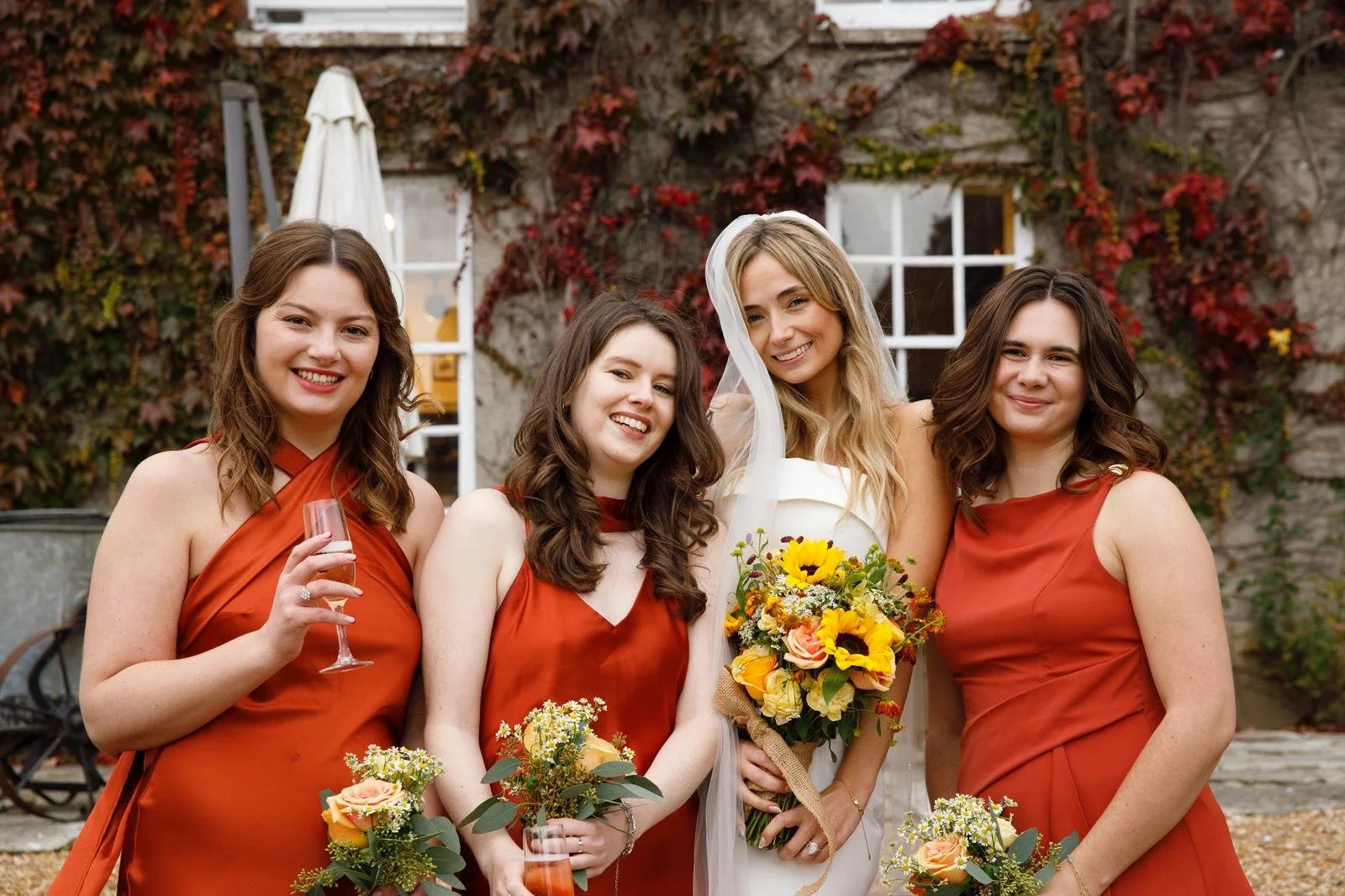 Four women dressed in autumn-colored dresses standing outdoors, smiling for a photo. One woman in the center is wearing a white wedding dress with a veil, holding a bouquet of sunflowers and other flowers. The other women are holding smaller bouquets