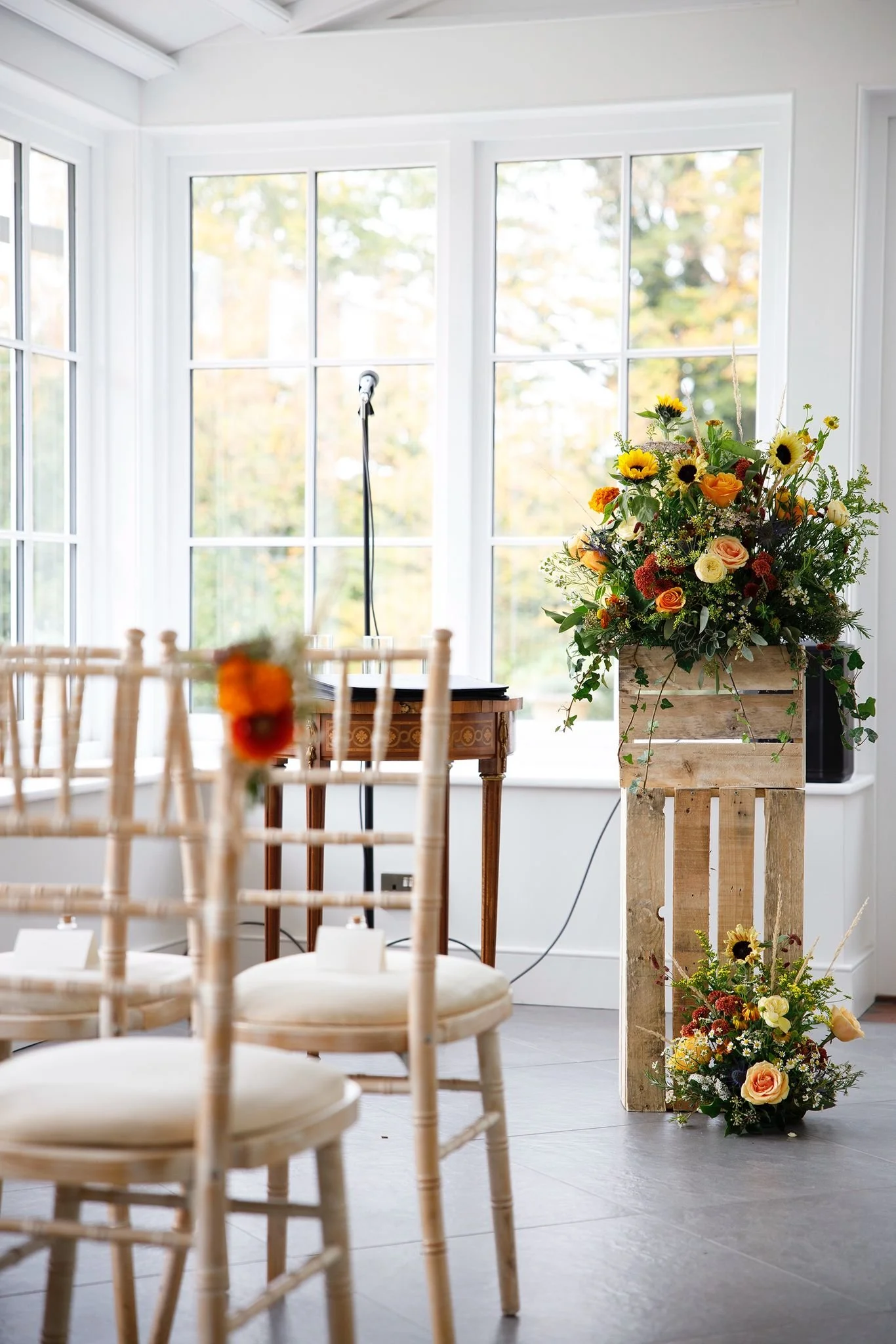 A decorated indoor space with a large floral arrangement on a wooden stand, a small table with a microphone, and multiple chairs arranged for an event or ceremony, with large windows showing trees outside.