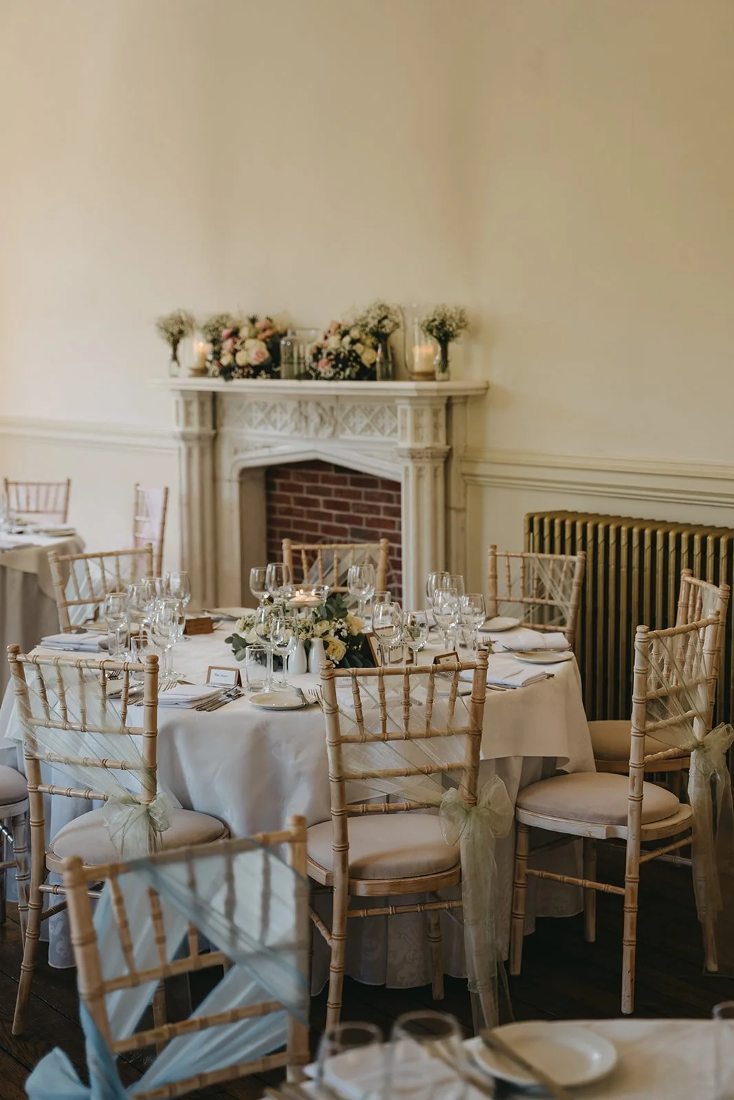Wedding reception table decorated with white tablecloth, floral centerpiece, and glassware, set in a room with a brick fireplace and candles, with chairs tied with light-colored ribbons.