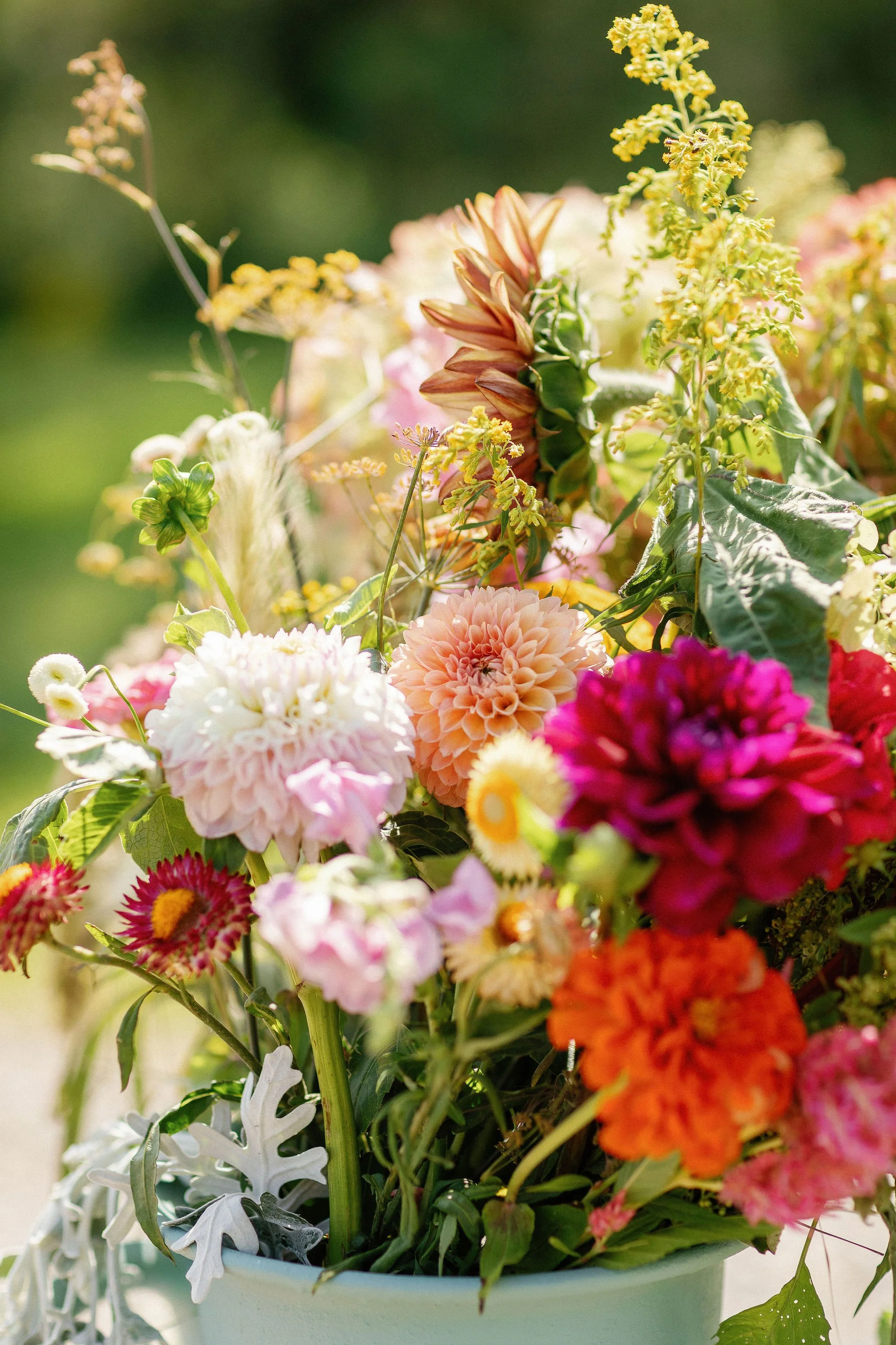 Colorful bouquet of various flowers, including dahlias and marigolds, in a white pot outdoors.