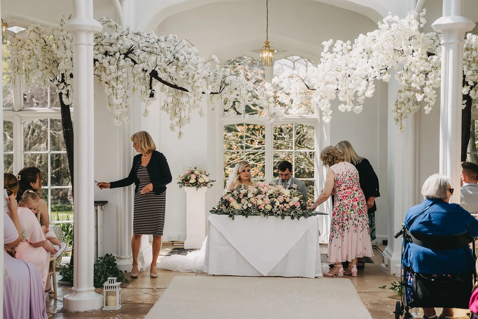 Indoor wedding ceremony with floral decor, including hanging white blossoms, with guests and bride and groom seated at a table.
