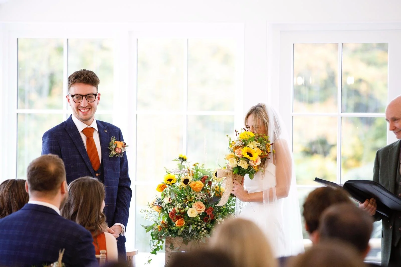 A wedding ceremony indoors with a bride presenting a bouquet of flowers and a groom smiling at guests.