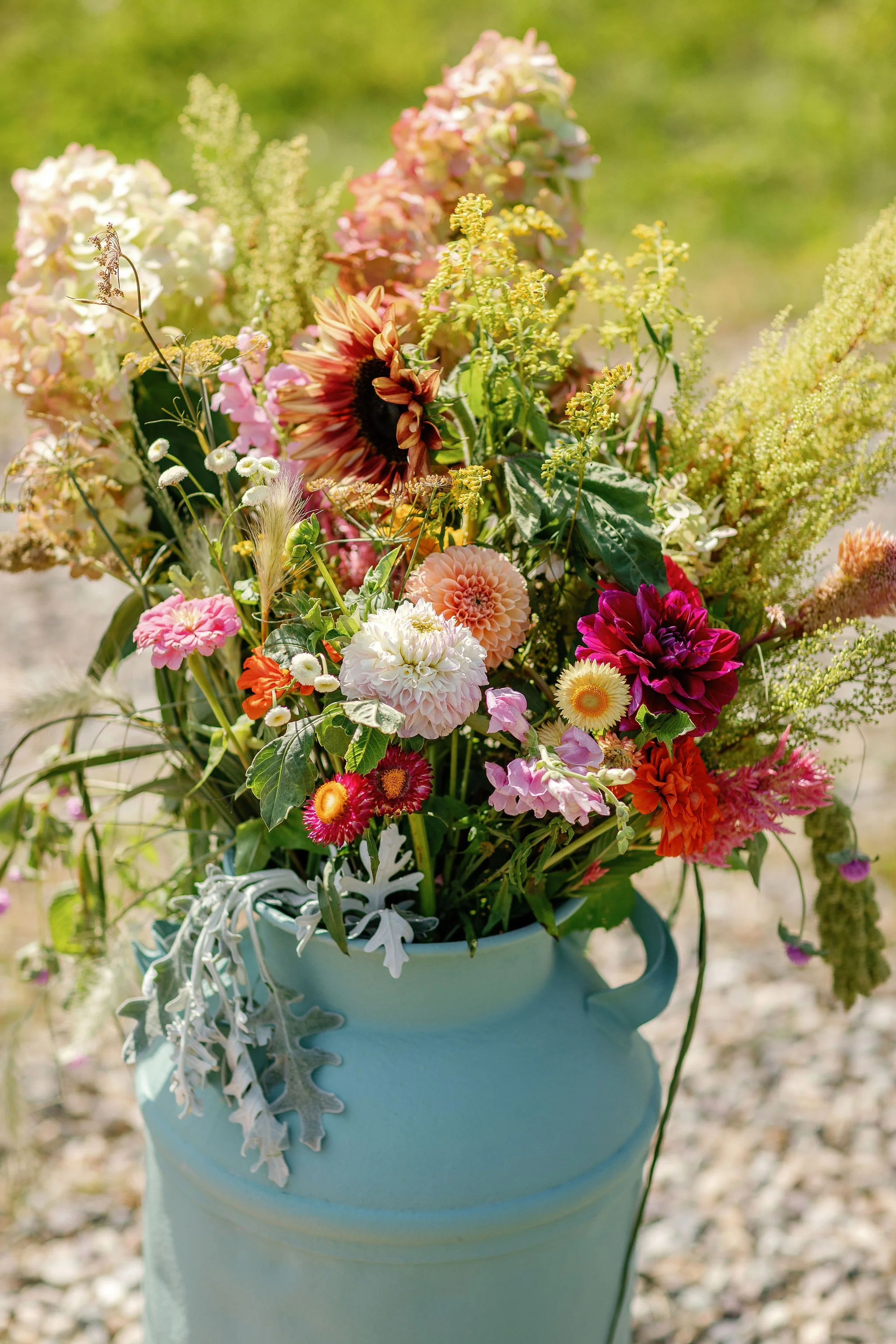 A bouquet of colorful flowers including dahlias, sunflowers, and other mixed blooms in a light blue vase outdoors.