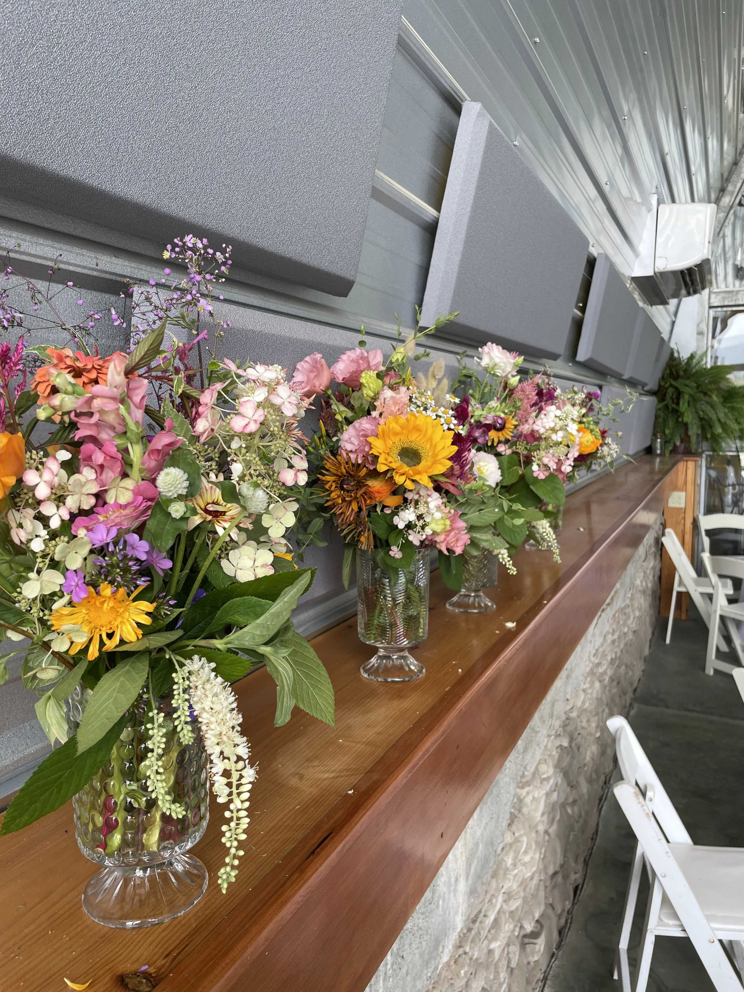 Various colorful flower arrangements in glass vases on a wooden ledge inside a building.