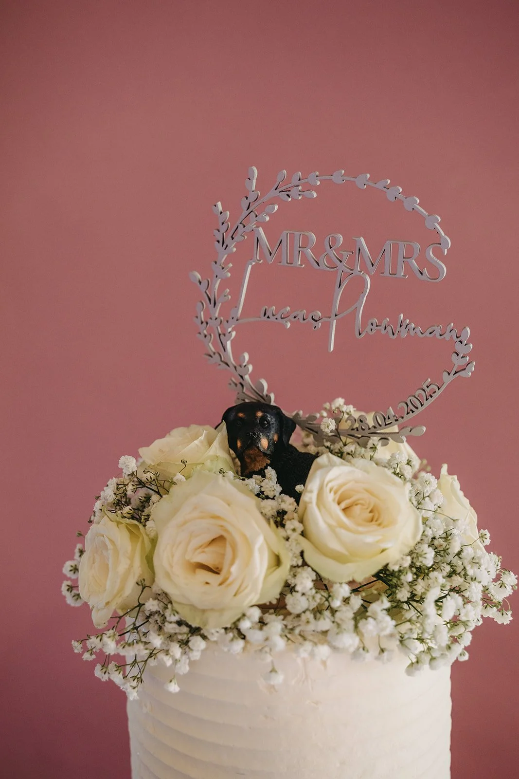 A wedding cake decorated with white roses and baby's breath flowers, topped with a sign that reads "Mr. & Mrs. Mac Aumann" and the wedding date "28.04.2023". A small black and brown dog peeks out from among the flowers against a pink background.