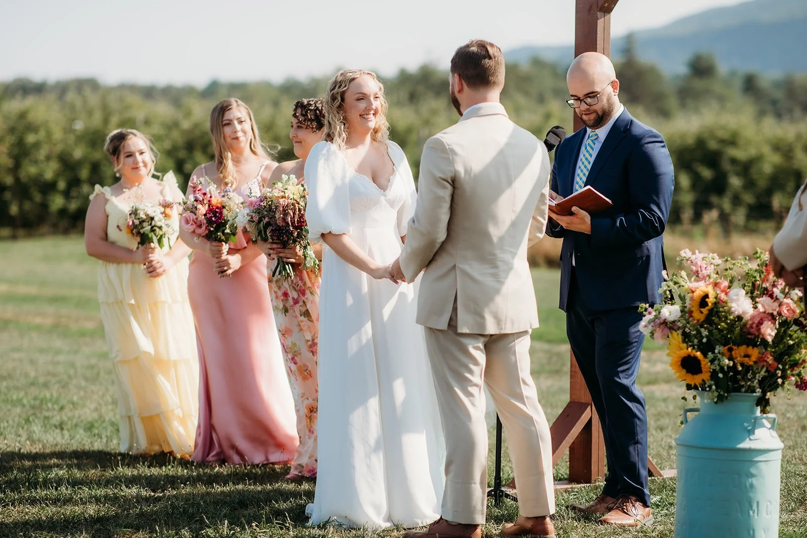 A couple is exchanging wedding vows outdoors, with an officiant standing nearby. Five bridesmaids holding bouquets are standing in line behind them, and a large blue container with colorful flowers is in the foreground, set against a backdrop of gree