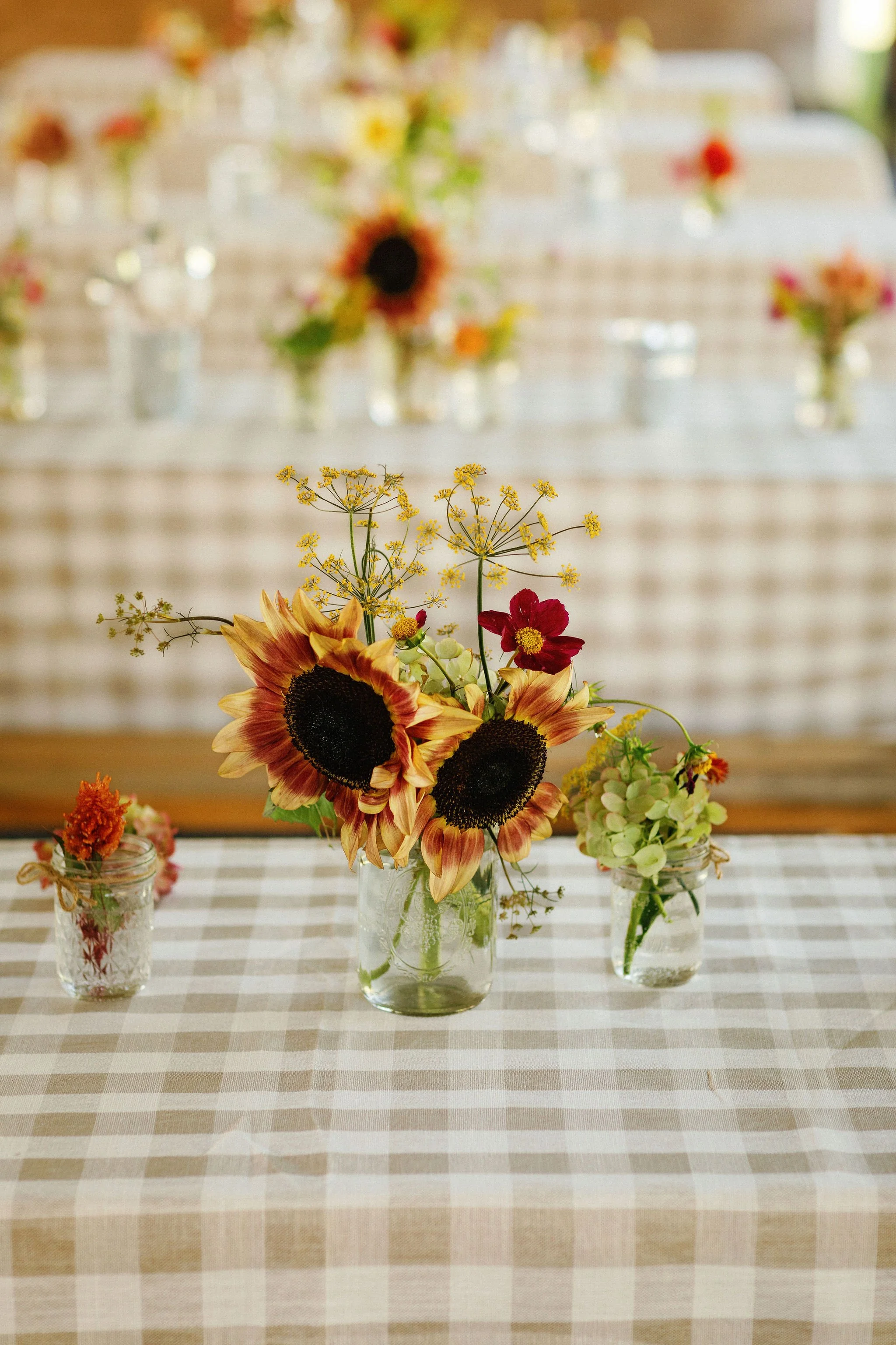 A flower centerpiece with sunflowers, red and cream-colored flowers, and yellow accents in a glass jar, flanked by smaller jars with orange, pink, and green flowers on a checkered tablecloth at a decorated event.