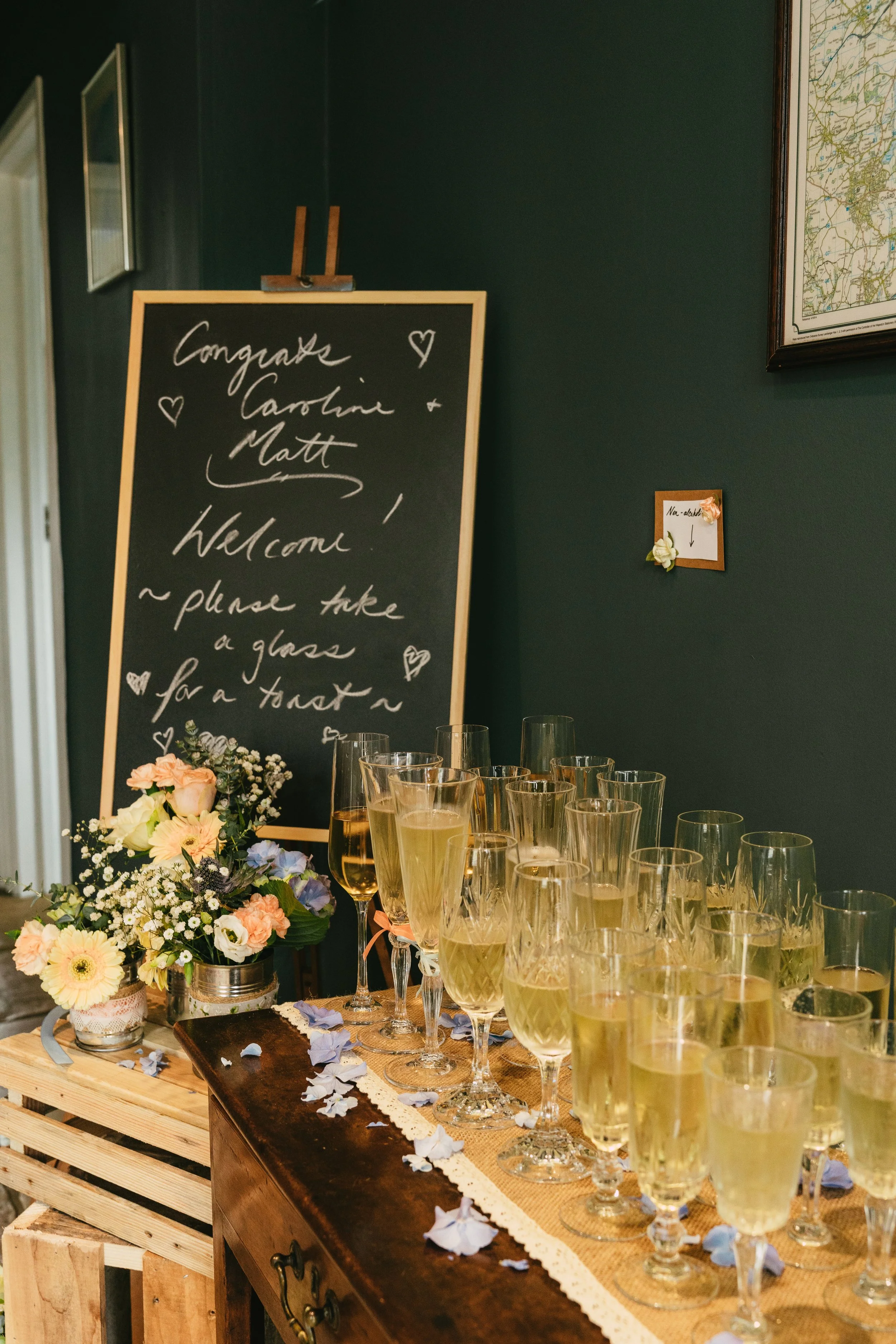 A table with multiple glasses of champagne beside a floral arrangement, a blackboard with handwritten message, and a small note on the wall.