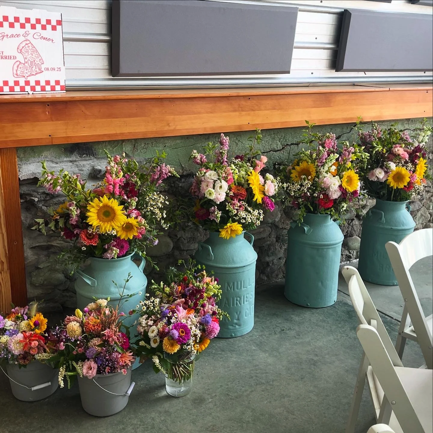 Five large blue vintage milk cans filled with colorful flowers, including sunflowers, pink, white, and purple blooms, are arranged along a stone wall on a concrete floor. A small arrangement of flowers in gray buckets and a glass vase are placed to t