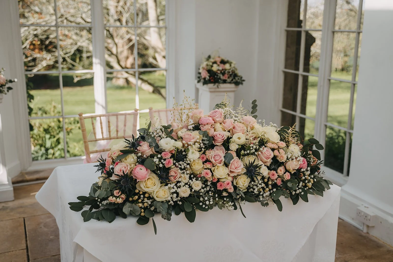A large floral arrangement of pink and white roses, baby's breath, and greenery on a white-covered table inside a sunroom with large windows showing a green garden outside.