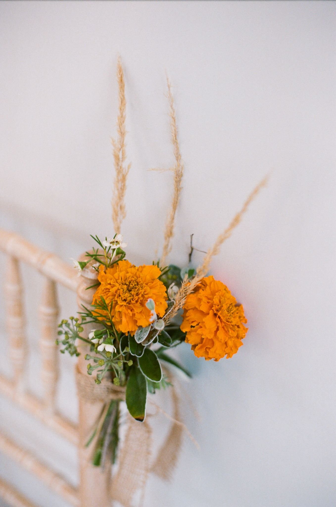 A small bouquet of yellow marigold flowers, white accent flowers, green leaves, and tall beige grass stems is attached to a light-colored wooden chair, set against a plain white wall.