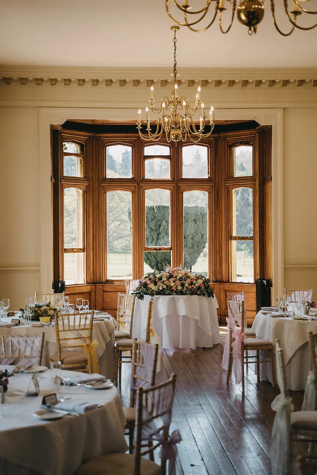 Elegant dining room set up for a wedding or event, with round tables covered in white tablecloths, surrounded by chairs decorated with pink ribbons, and a floral centerpiece on a table in front of a large bay window with wooden trim, natural light co