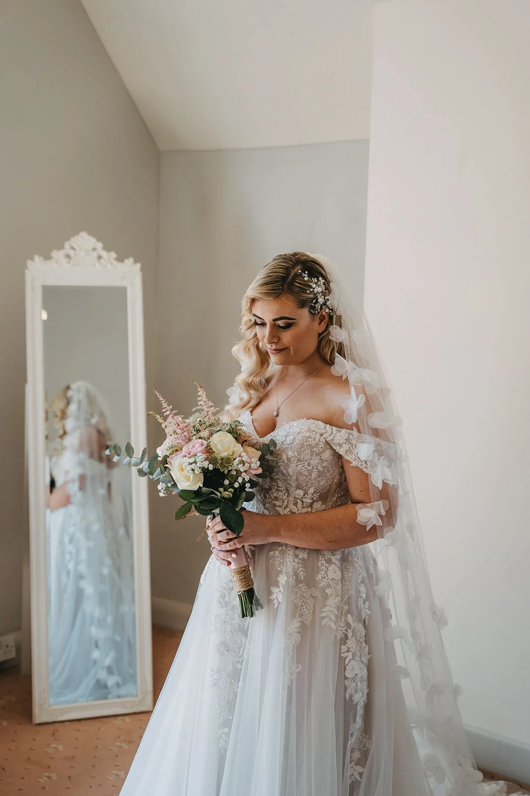 A bride in a lace wedding gown holding a bouquet of white and pink flowers, standing in front of a mirror with an ornate white frame, inside a room with beige walls and a beige carpet.