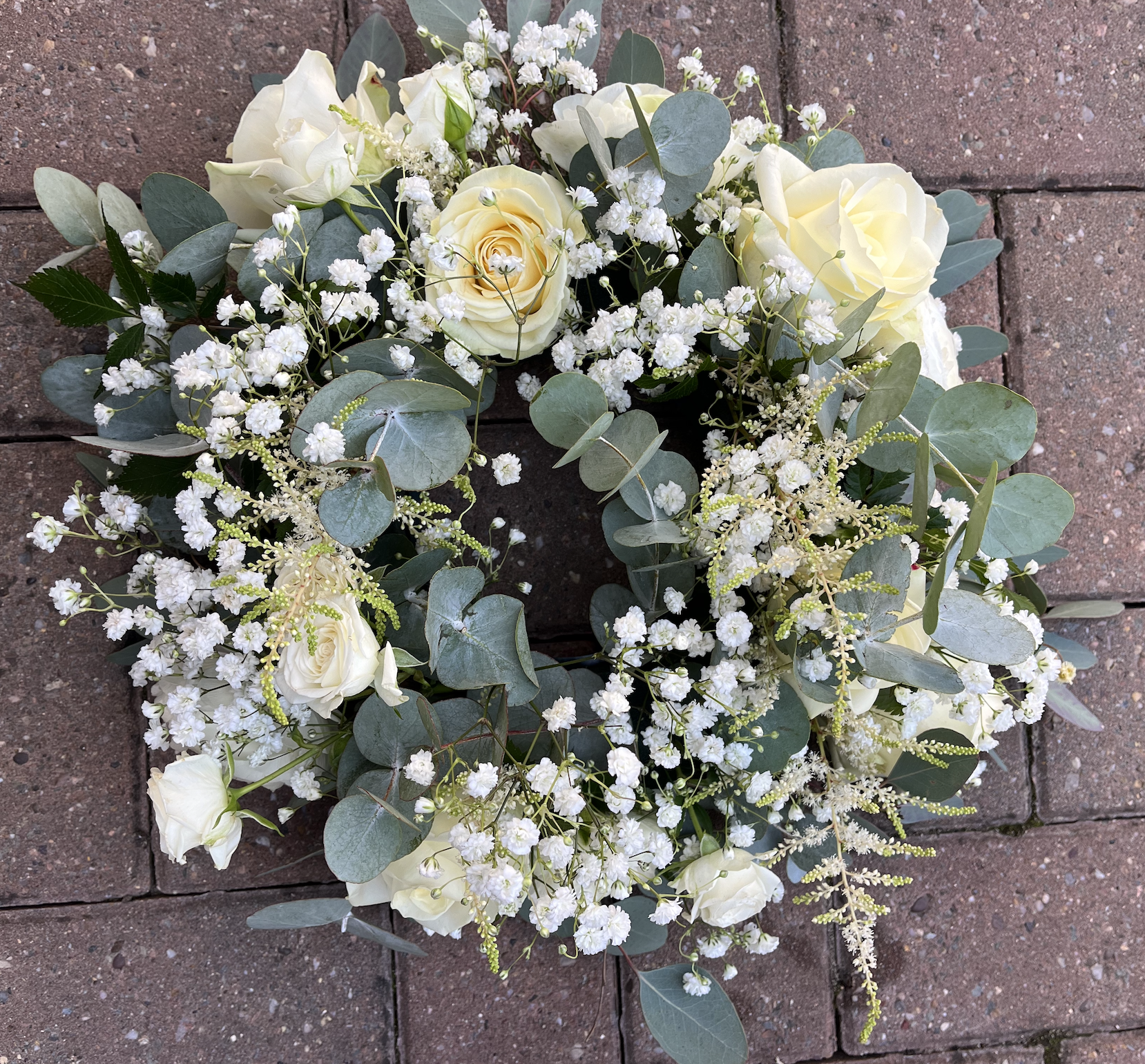 A white floral wreath with white roses, baby's breath, and eucalyptus leaves on a brick surface.
