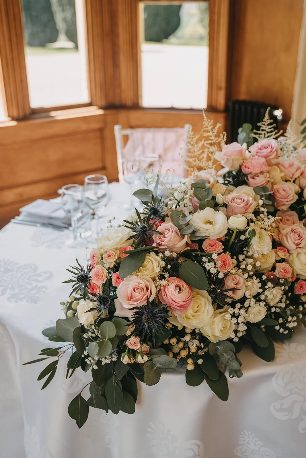 A floral centerpiece on a white tablecloth at a wedding reception, with pink and white roses, eucalyptus leaves, and dark thistle flowers, set in a wooden-paneled room with windows.