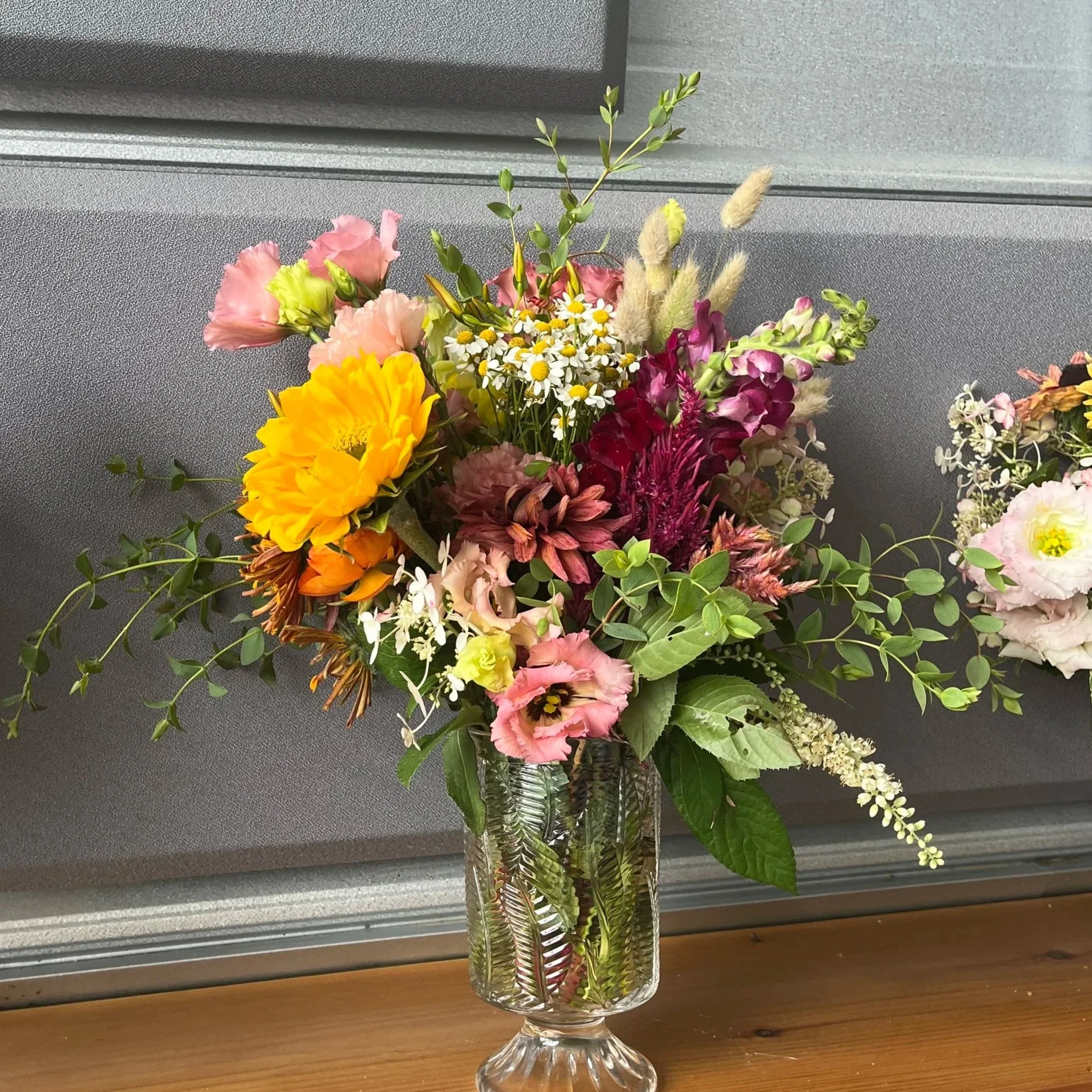 Colorful bouquet of mixed flowers in a tall glass vase on a wooden surface, with a gray wall in the background.