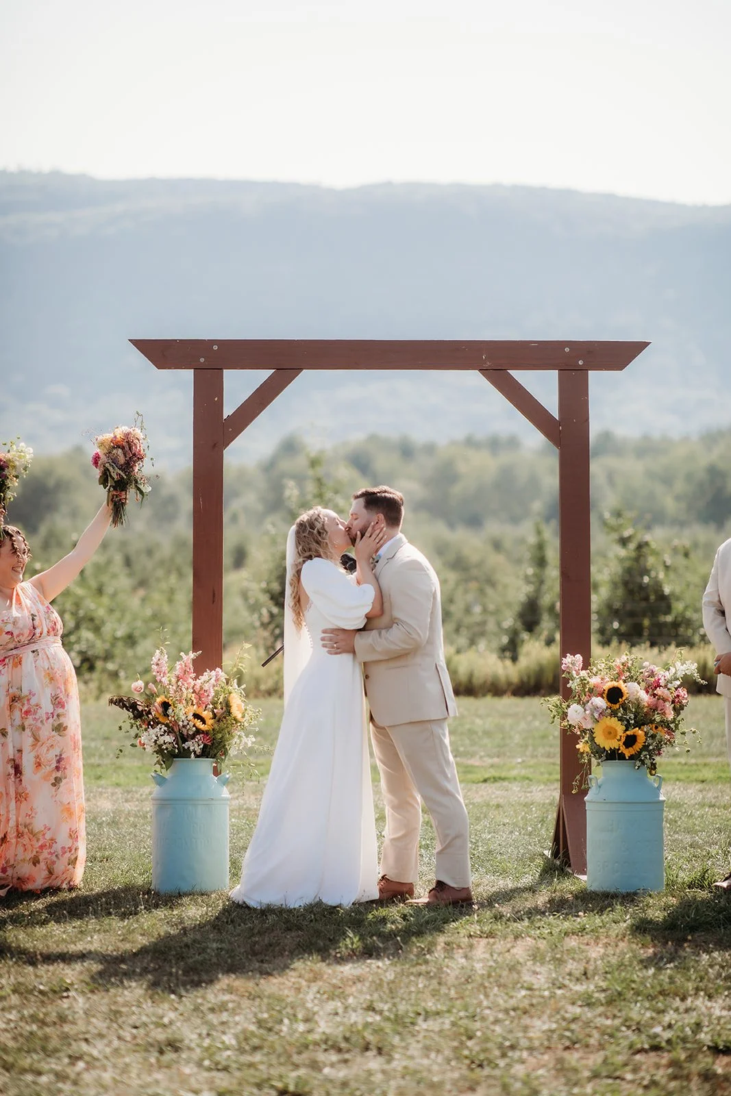 A couple kiss during an outdoor wedding ceremony under a wooden arch, with two women holding flower bouquets nearby and a scenic mountain background.