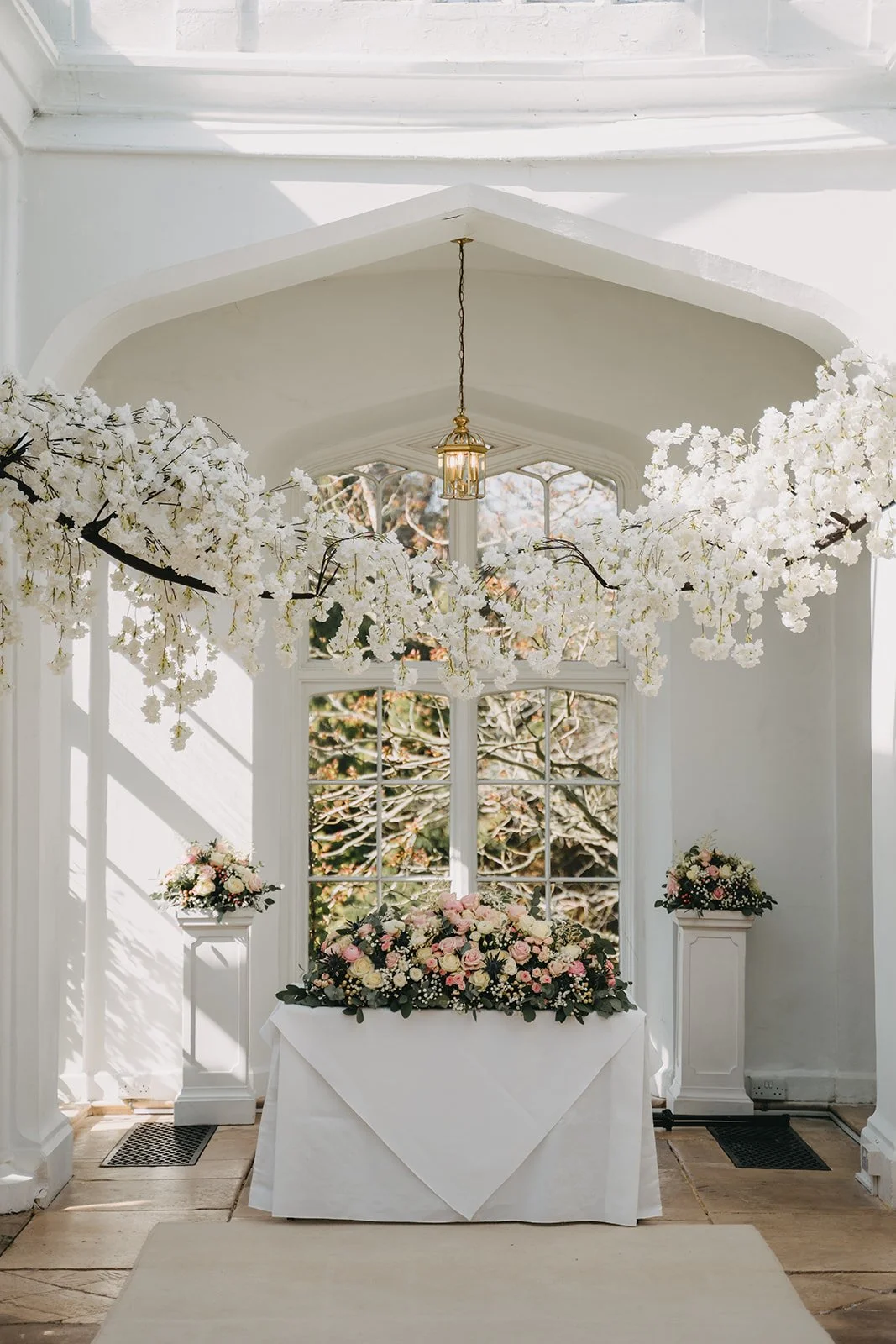 Interior of a bright room decorated for a wedding, with white floral arrangements, a table with a large floral centerpiece, and windows showing trees outside.