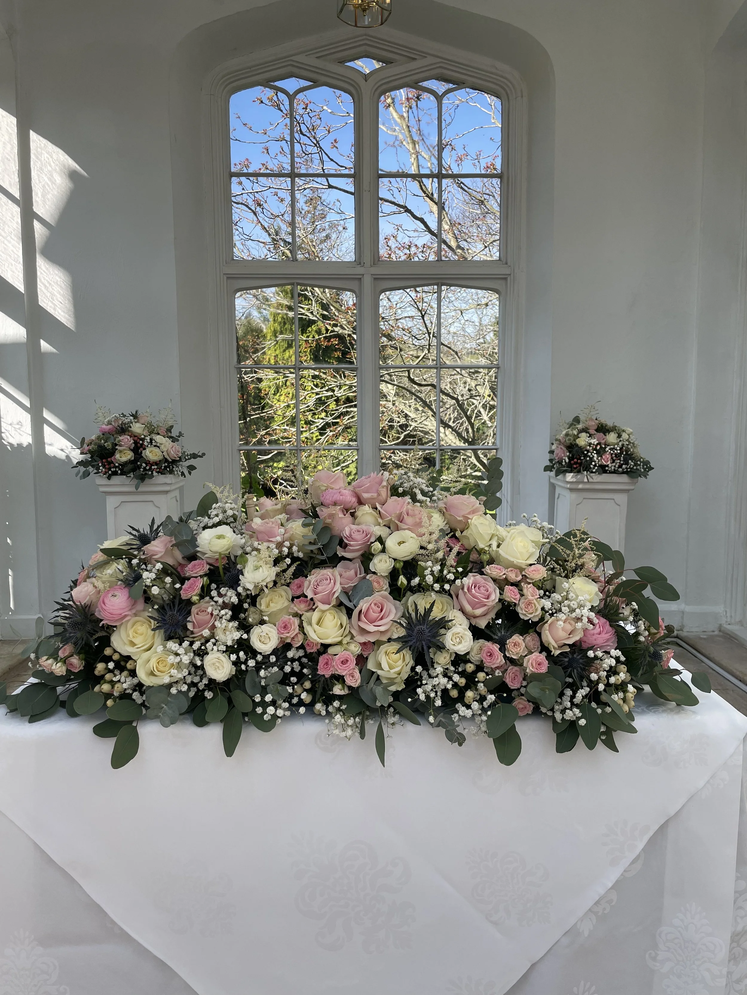A floral arrangement of pink, white, and cream roses, baby's breath, and greenery on a table with a white tablecloth, set in front of a large window with an outdoor view of trees.
