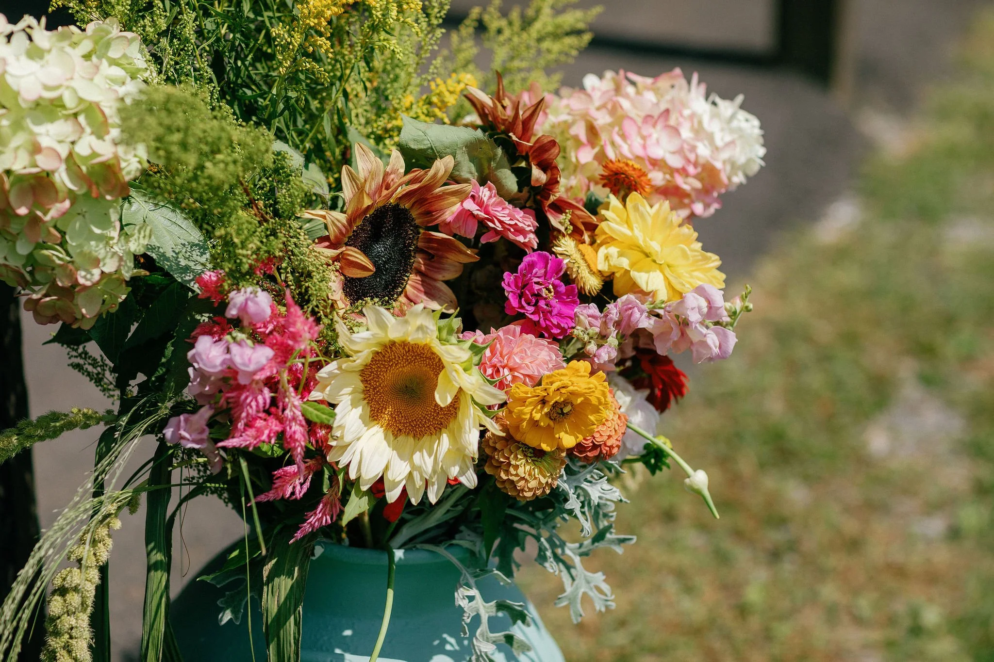Colorful bouquet of various flowers, including sunflowers, roses, and hydrangeas, arranged in a vase outdoors.