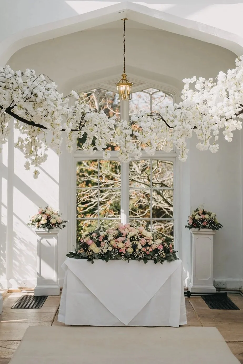 A decorated indoor space with white walls and a large window showing trees outside. The ceiling has white floral arrangements hanging from a gold chandelier. A table covered with a white cloth displays a large pink and white flower arrangement, flanked by two smaller flower arrangements on white pedestals.