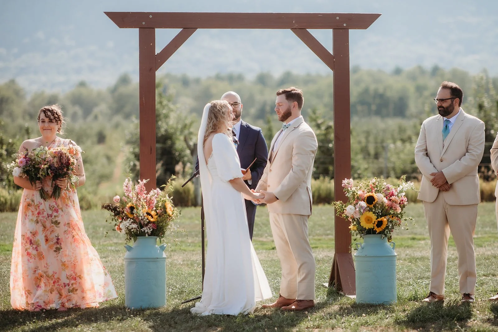 A couple exchanging vows at an outdoor wedding ceremony with an officiant, two bridesmaids, and floral decorations on a sunny day.