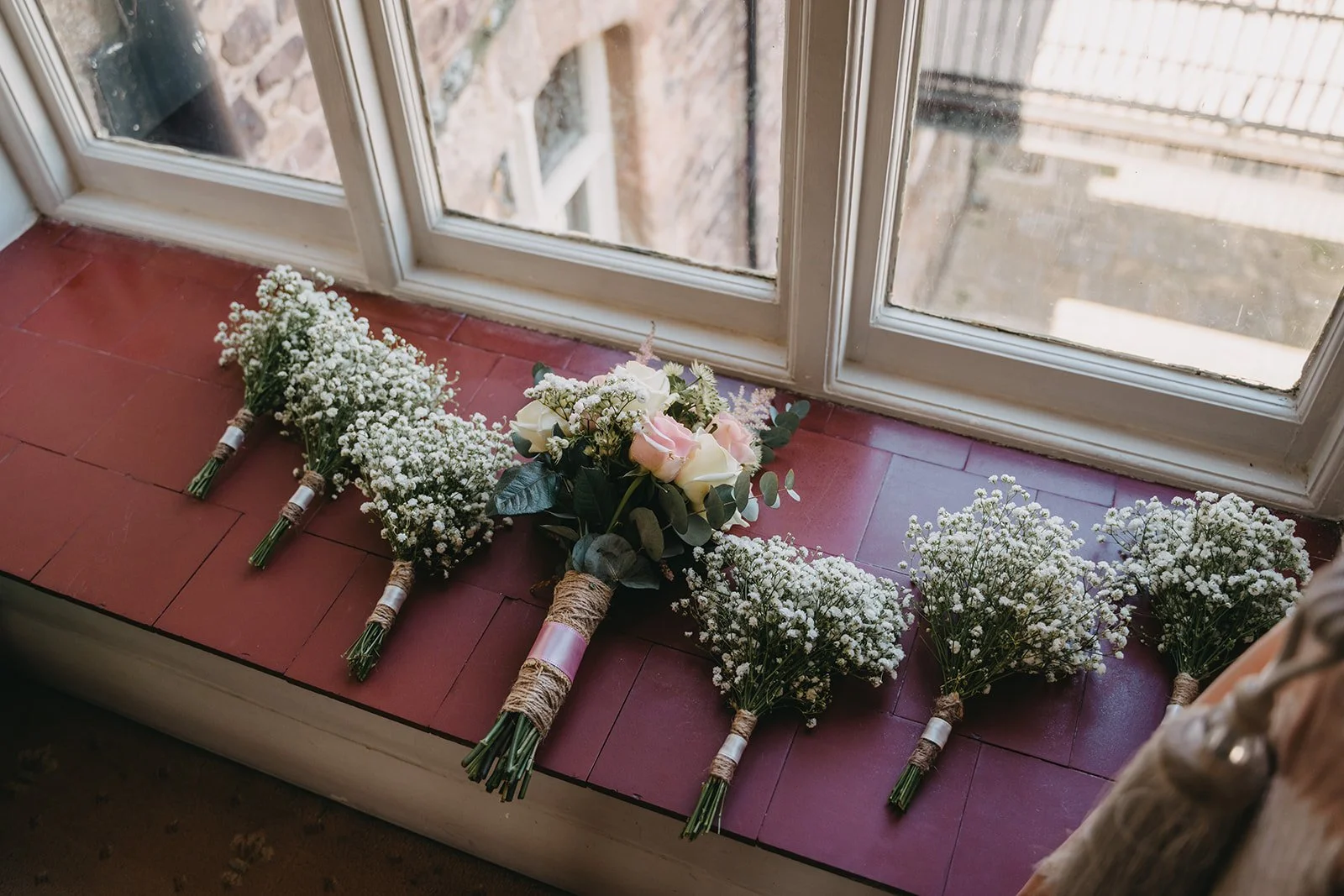 Five small bouquets of white baby's breath flowers and one large bouquet of white and pink roses with greenery, laid out on a windowsill with a red tiled surface.